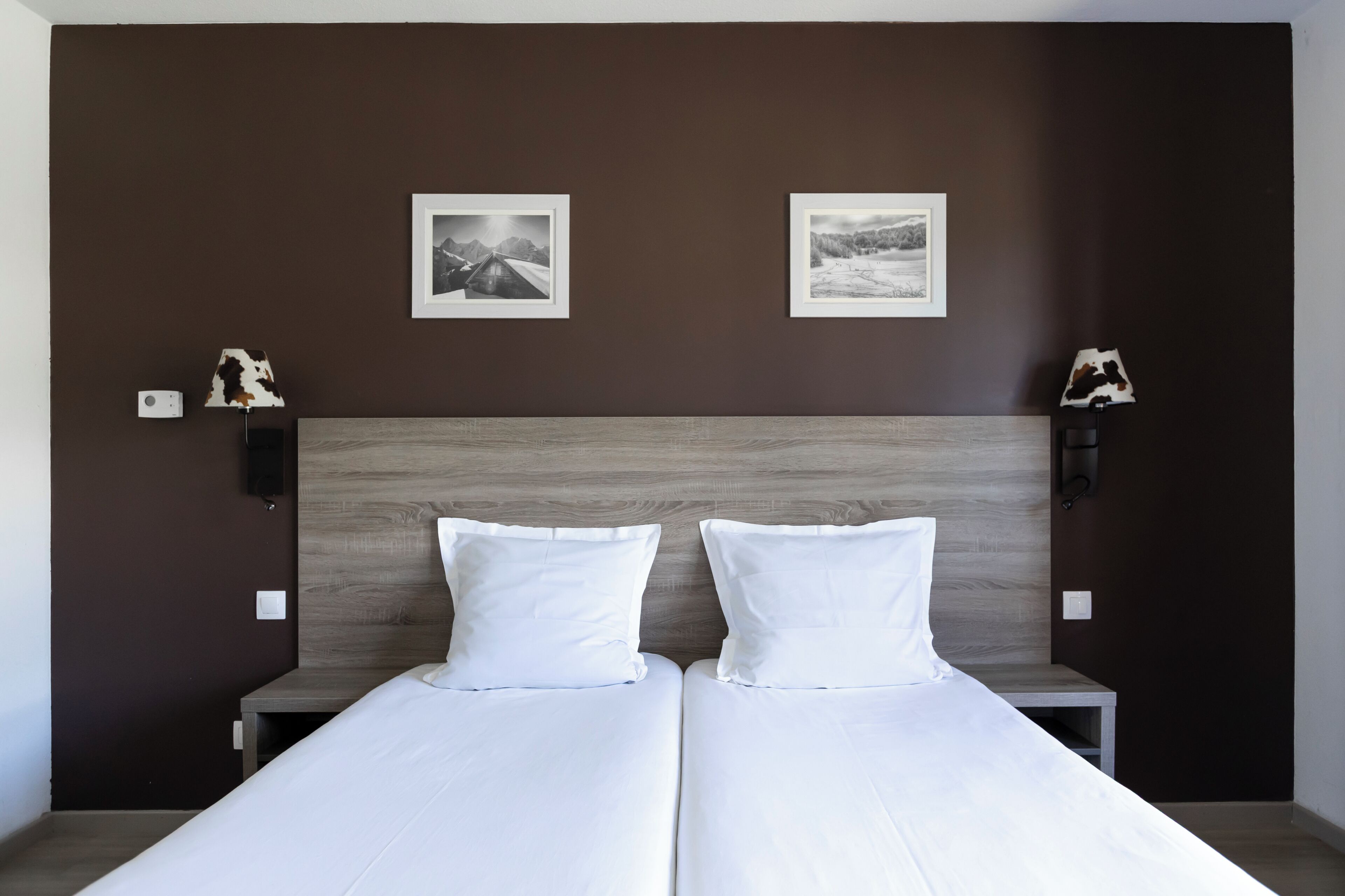 Cama doble con ropa de cama blanca, cabecera de madera, dos cuadros enmarcados en la pared y una mesita de noche con una lámpara.