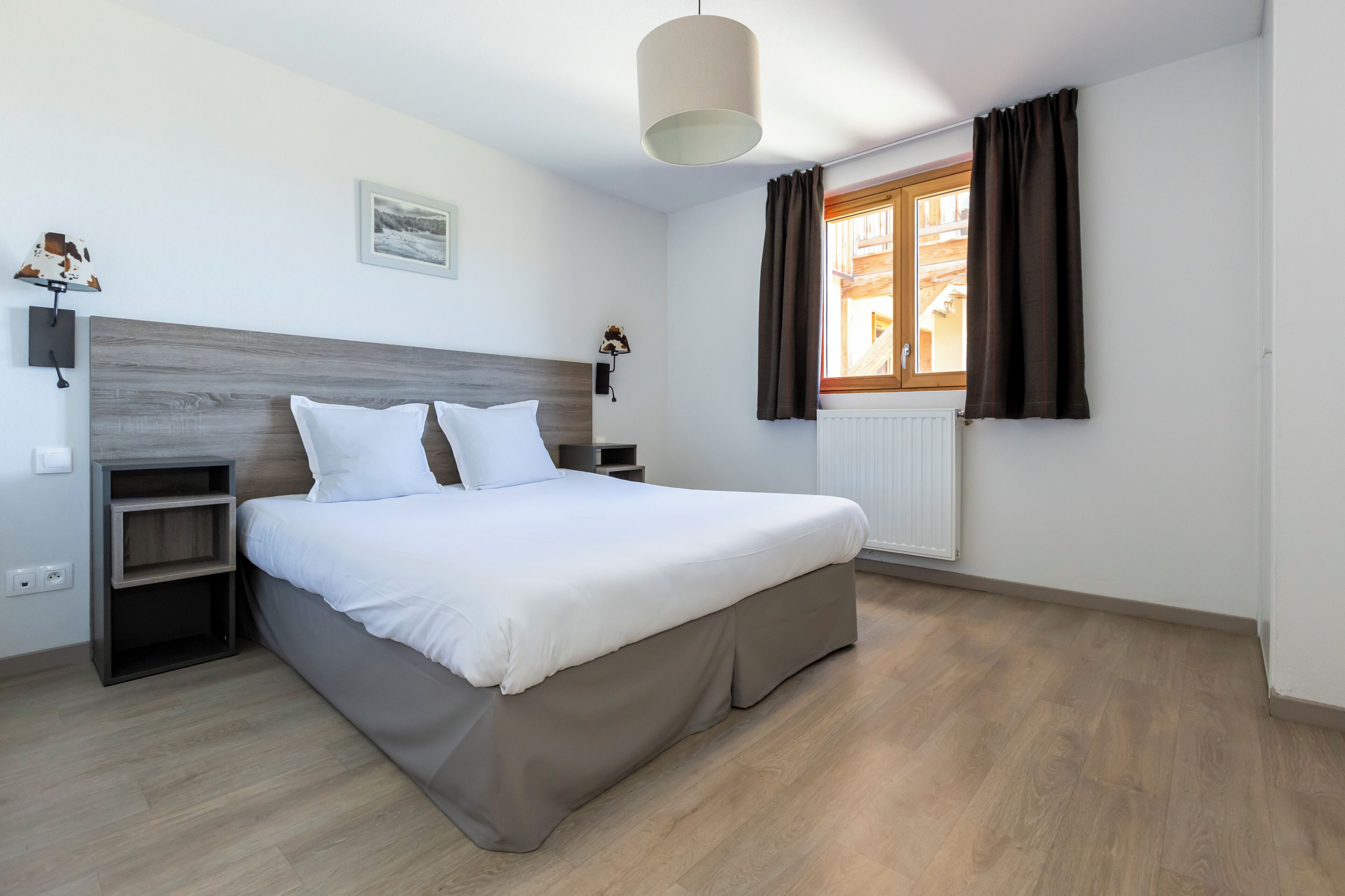 Une chambre à coucher avec un lit, des tables de chevet, une lampe, une fenêtre avec des rideaux et un radiateur.