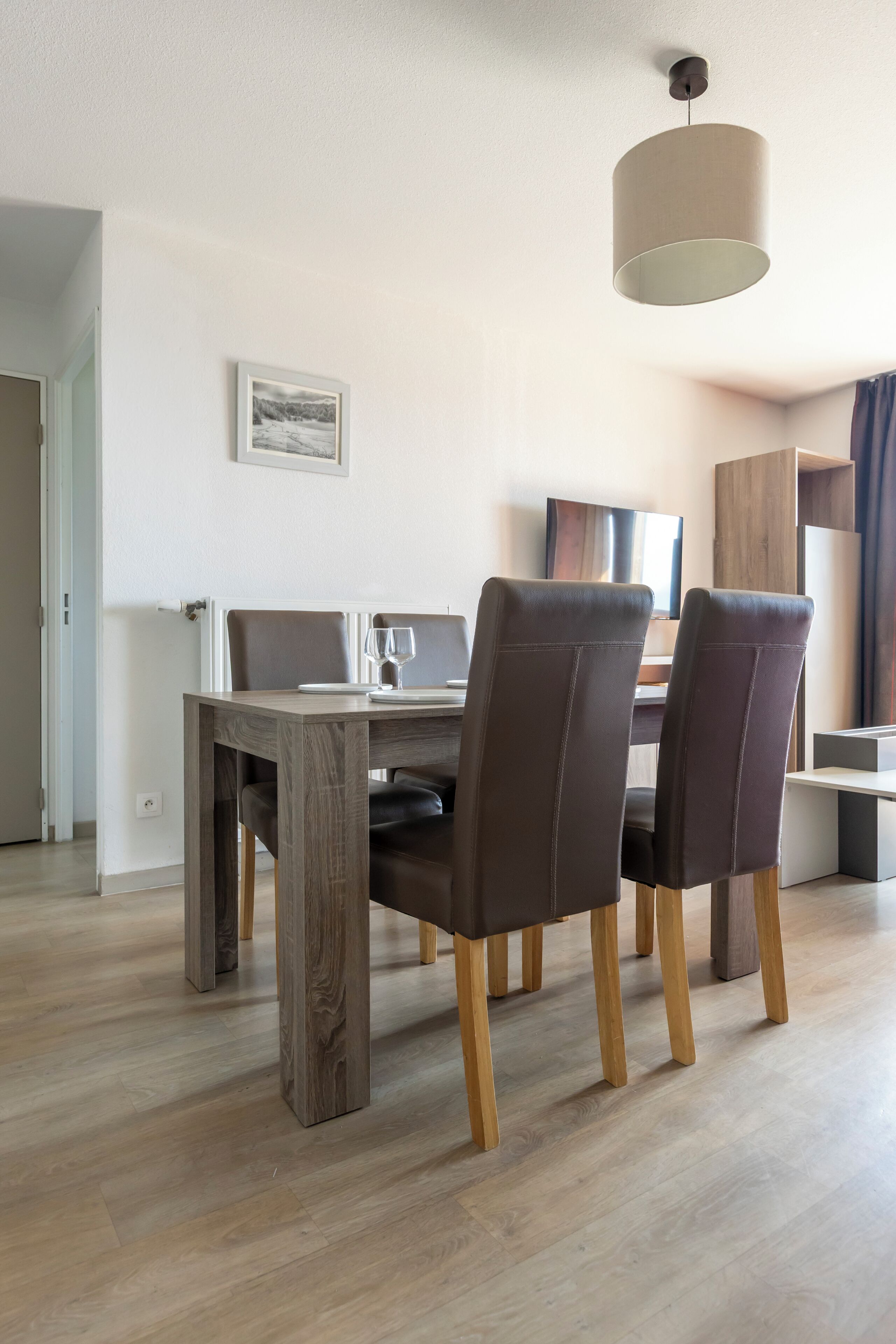 A dining area with a wooden table, two chairs, a hanging light fixture, and a framed picture on the wall.