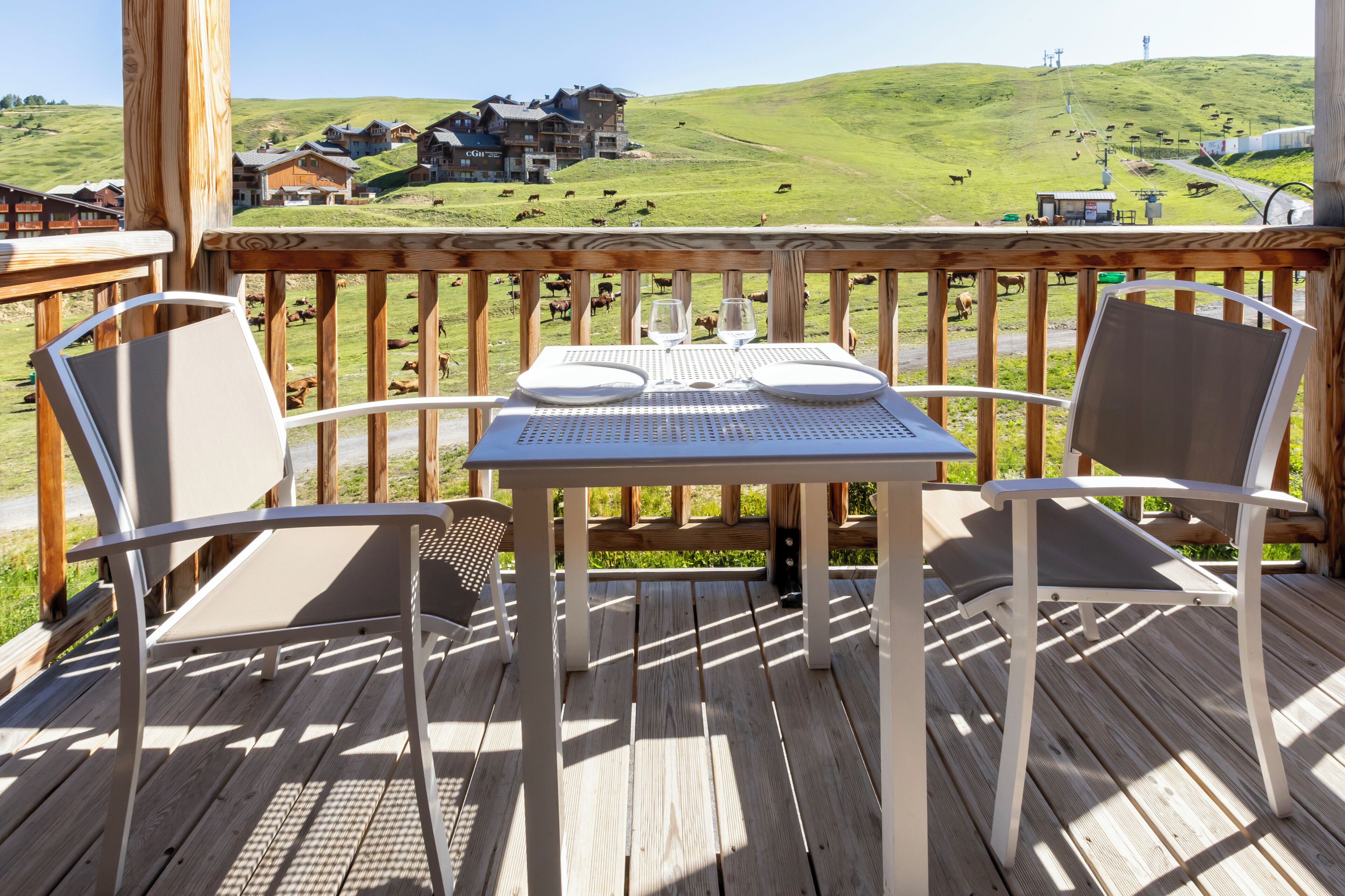 A wooden deck with a table and chairs, overlooking a green landscape with buildings and a ski lift.