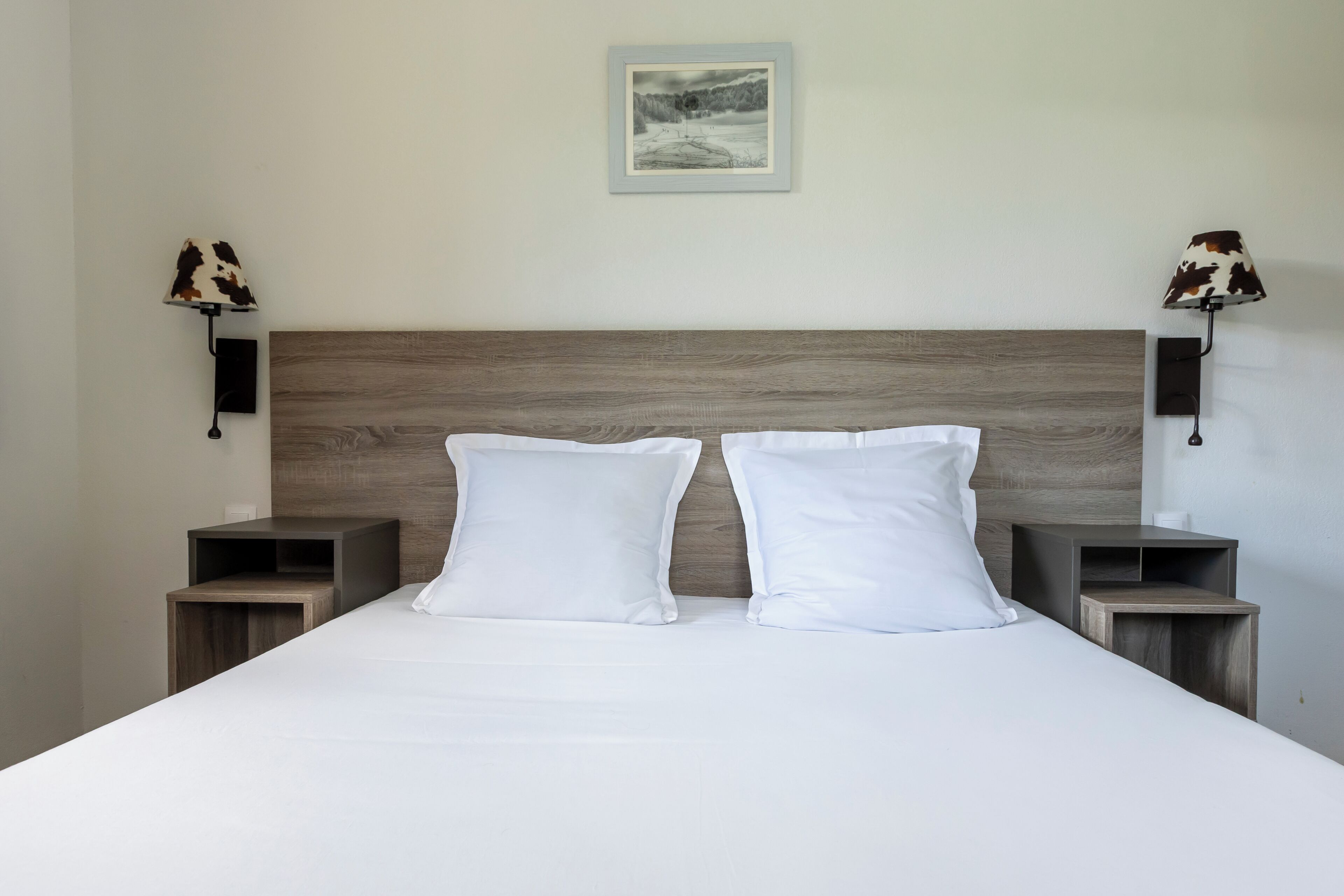 A neatly made bed with white linens and two pillows, flanked by two bedside tables with lamps, in a room with a wooden headboard and a framed picture on the wall.