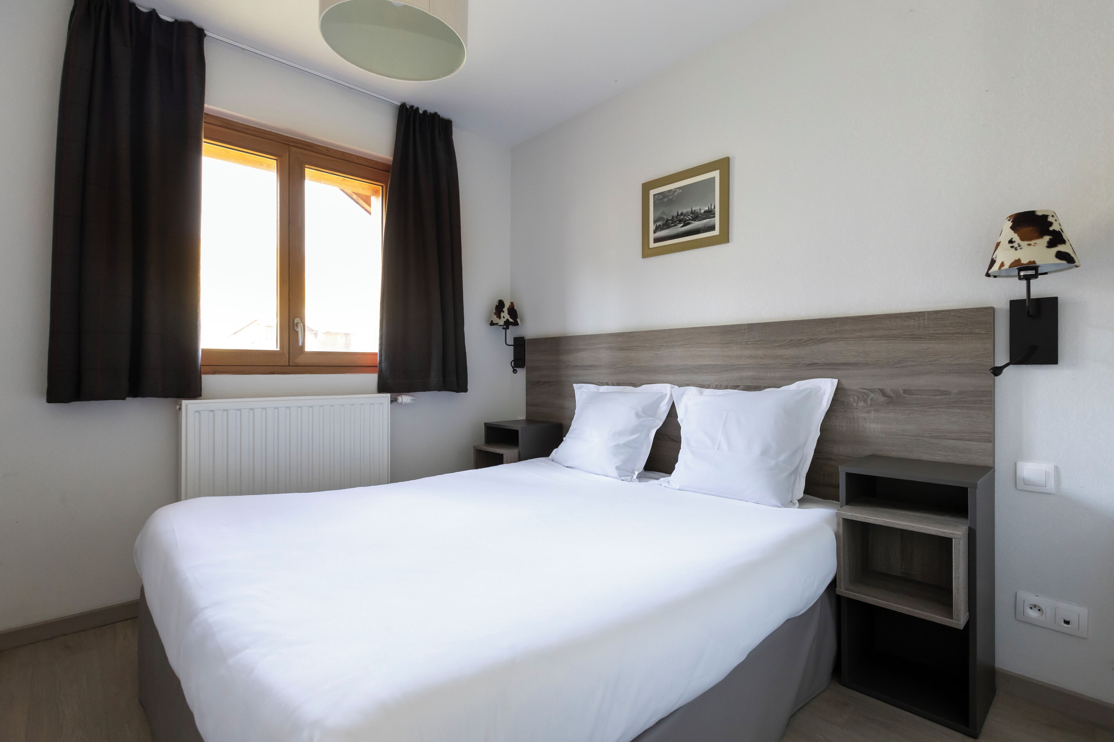 Una cama bien hecha con sábanas blancas, cabecera de madera, mesita de noche con lámpara y ventana con cortinas.