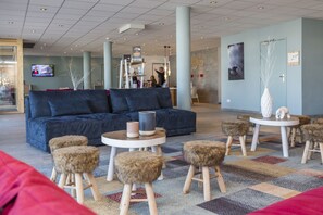 Un hall modern amb un sofà blau fosc, taules rodones amb tamborets i una televisió que mostra un programa d'informatius.