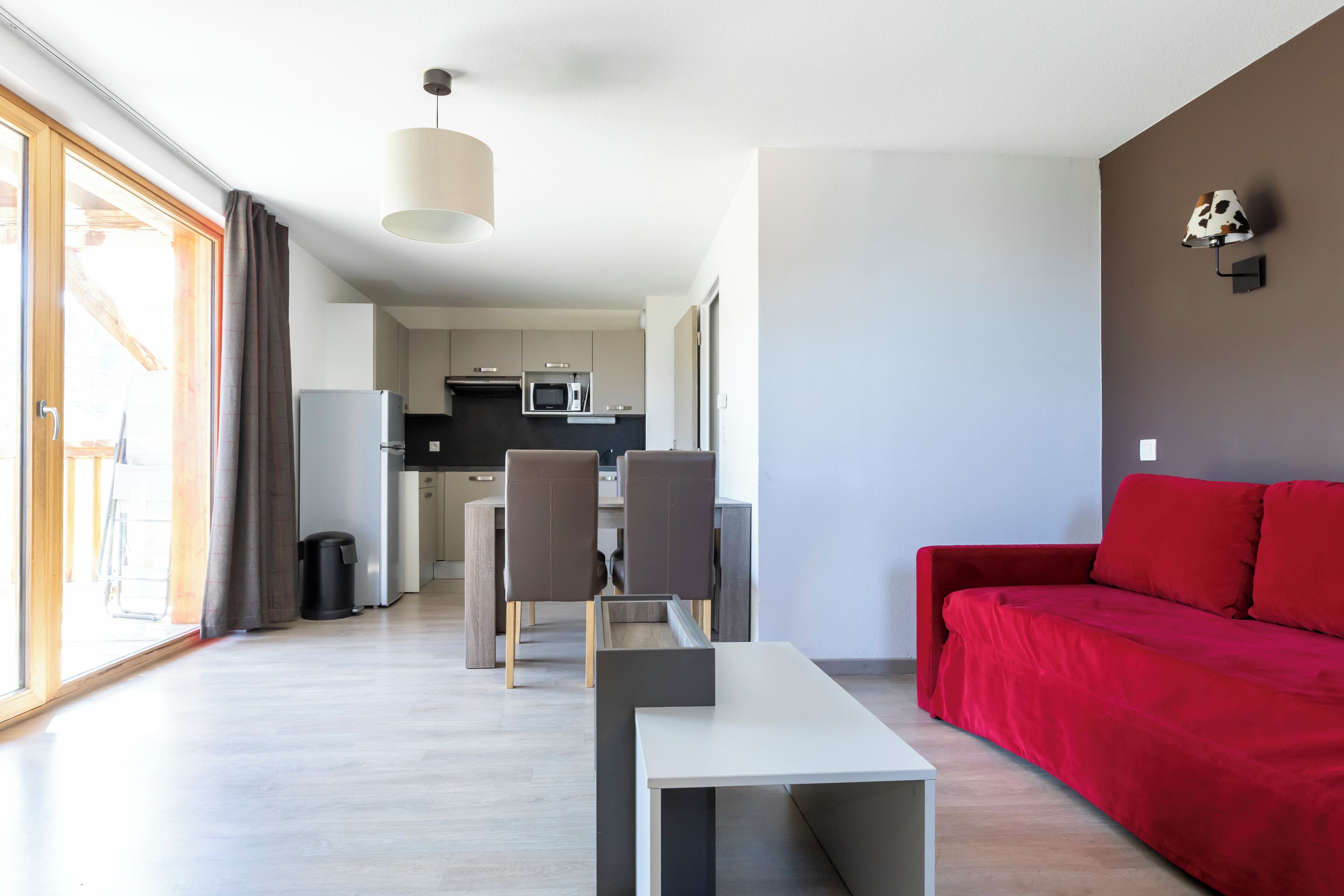 Una sala de estar moderna con un sofá rojo, un área de comedor con sillas, una cocina al fondo y un ventanal grande.