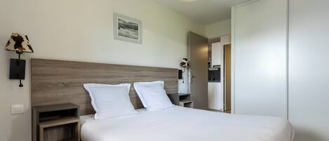 A hotel room with a wooden headboard, a bed with white linens, a nightstand, a wall-mounted lamp, and a framed picture.