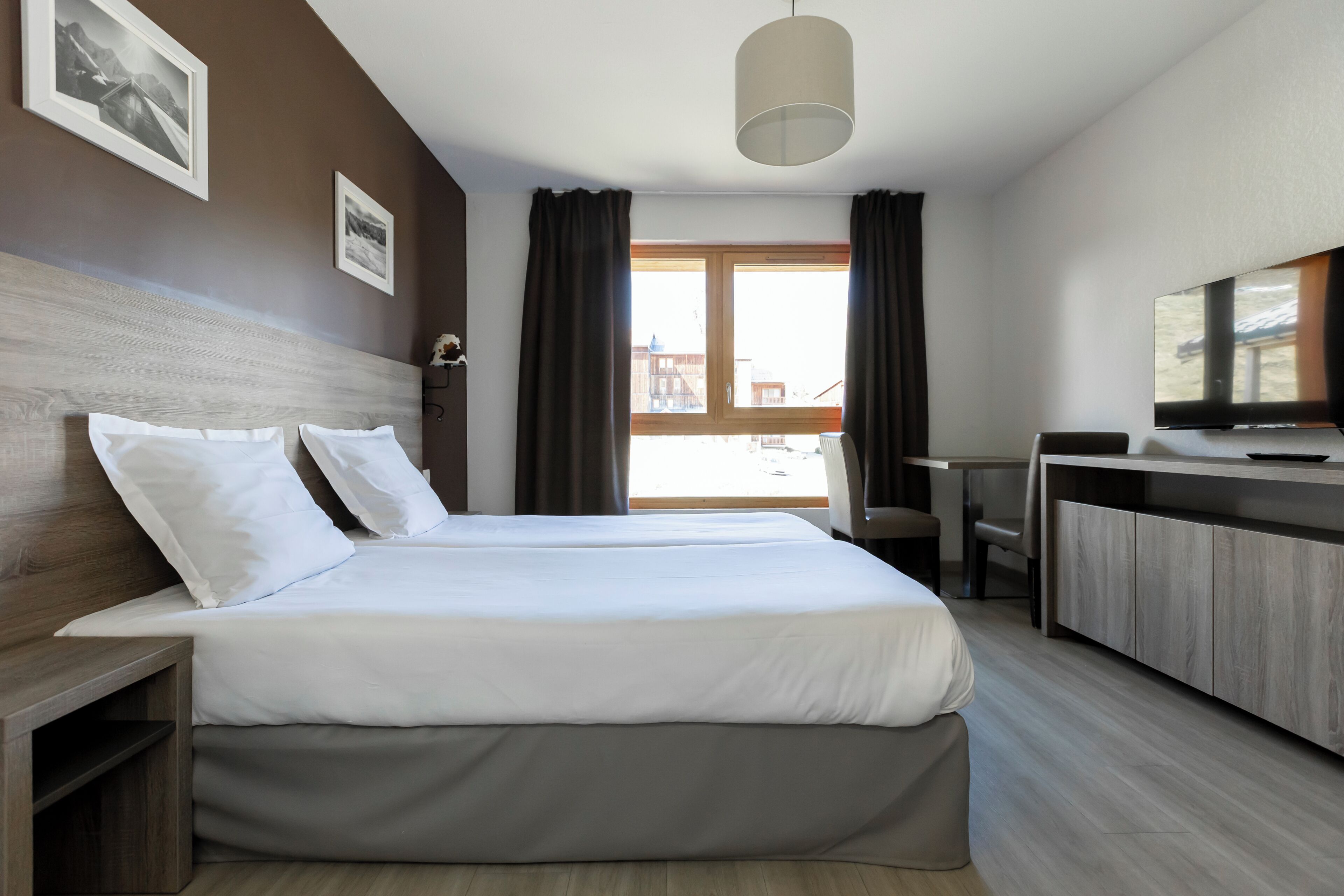 Habitación de hotel con dos camas, un escritorio, una silla, televisión y ventana con cortinas.