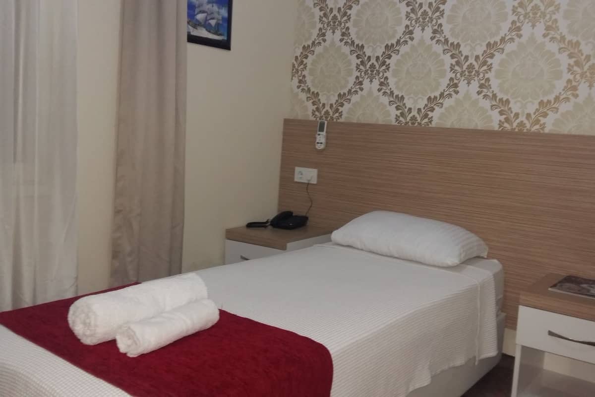 Una cama bien hecha con una colcha roja y dos toallas enrolladas, una mesita de noche con un teléfono y una revista, y una pared con papel tapiz estampado.
