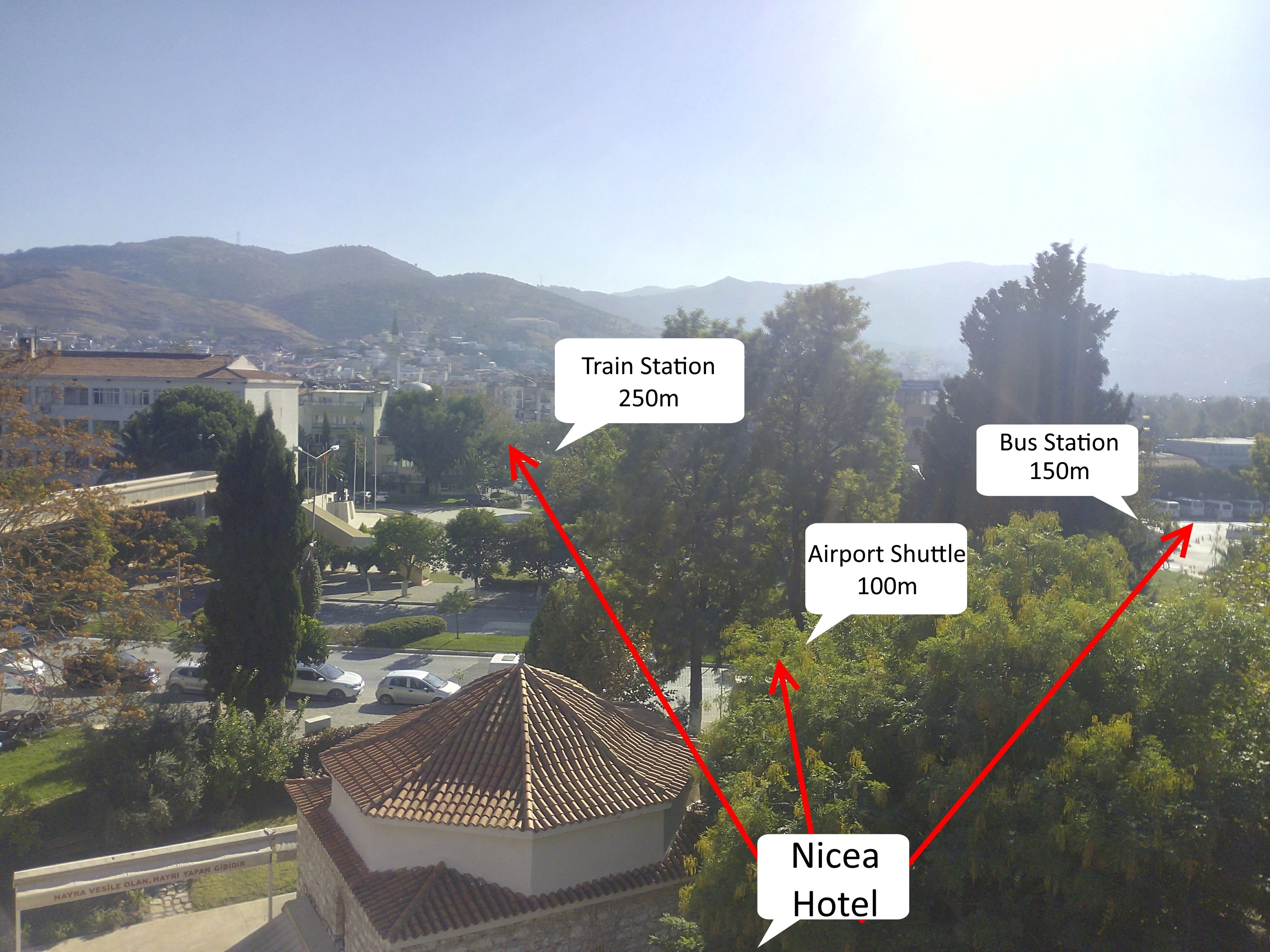Vista de un hotel con techo de tejas, rodeado de vegetación y un paisaje urbano al fondo.