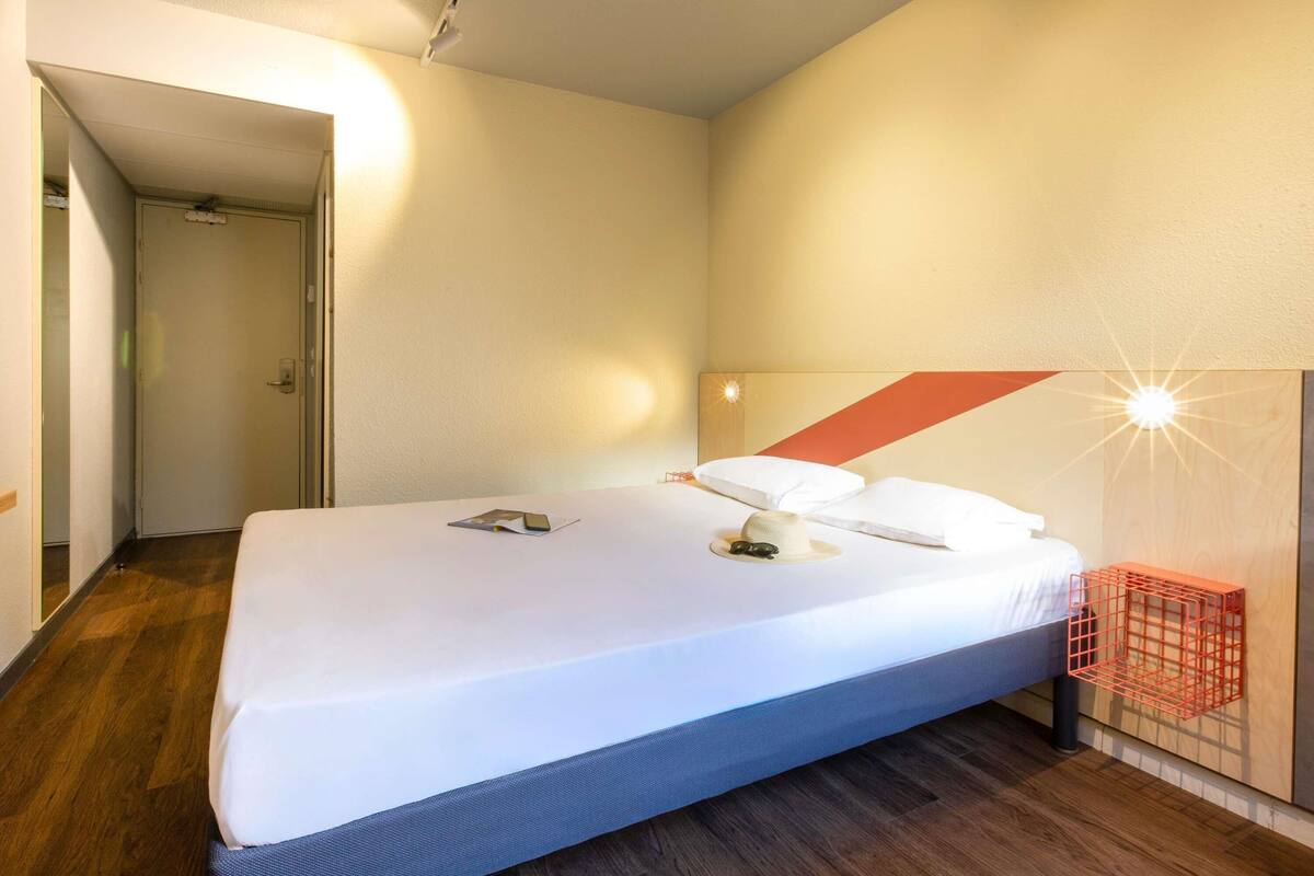 A modern hotel room with a large bed, a wooden floor, a door, and a wall-mounted light fixture.
