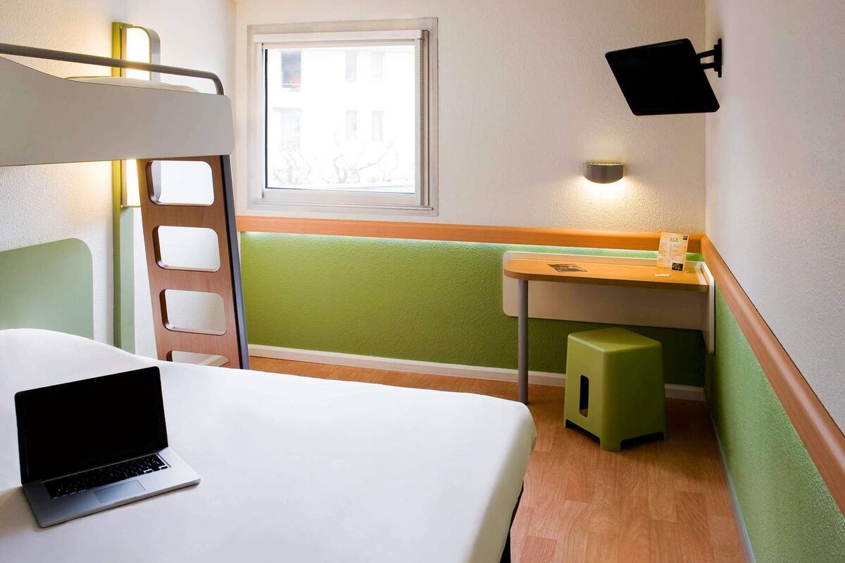 A hotel room with a bunk bed, a desk with a chair, a laptop, a window with a view, and a wall-mounted lamp.
