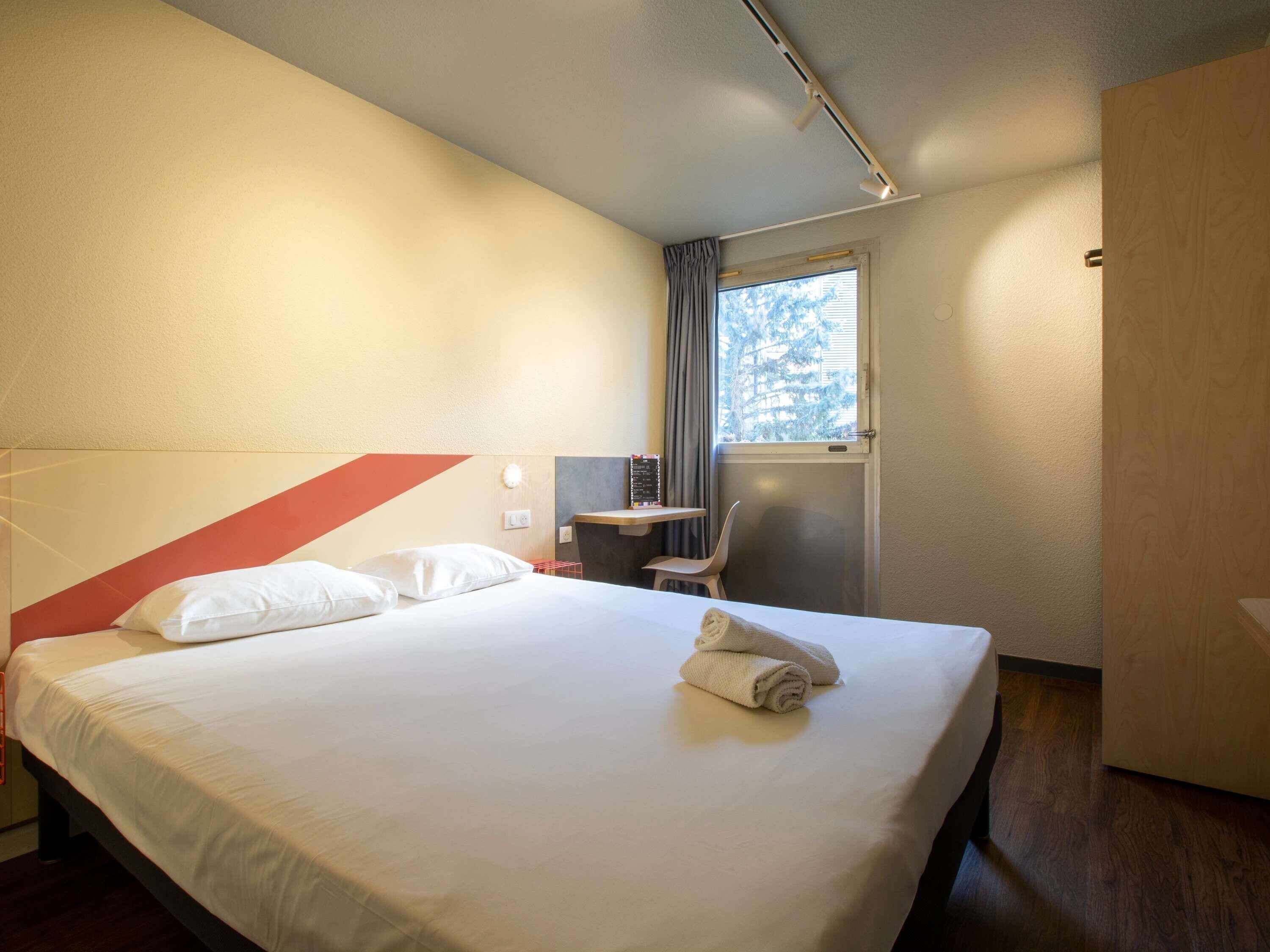 A hotel room with a bed, a desk, a chair, a window, and a door.
