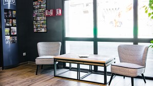 A modern room with a wooden floor, two patterned chairs, a small table, a bookshelf with magazines, and large windows.