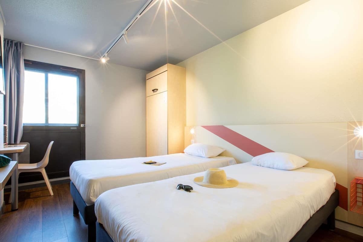 A hotel room with two beds, a desk, a chair, and a window.