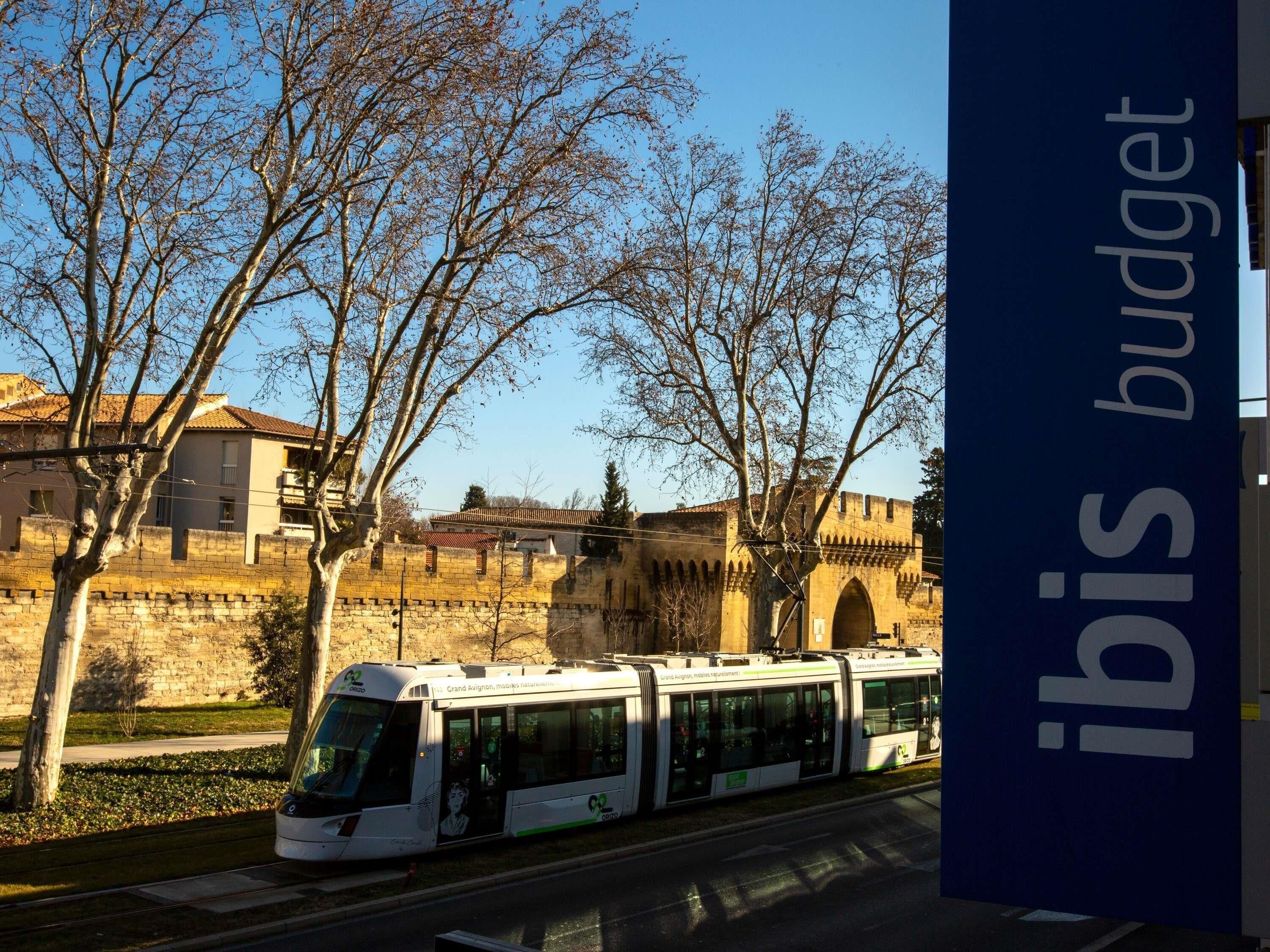 A tram with a white and green design, passing by a historic stone wall and leafless trees.