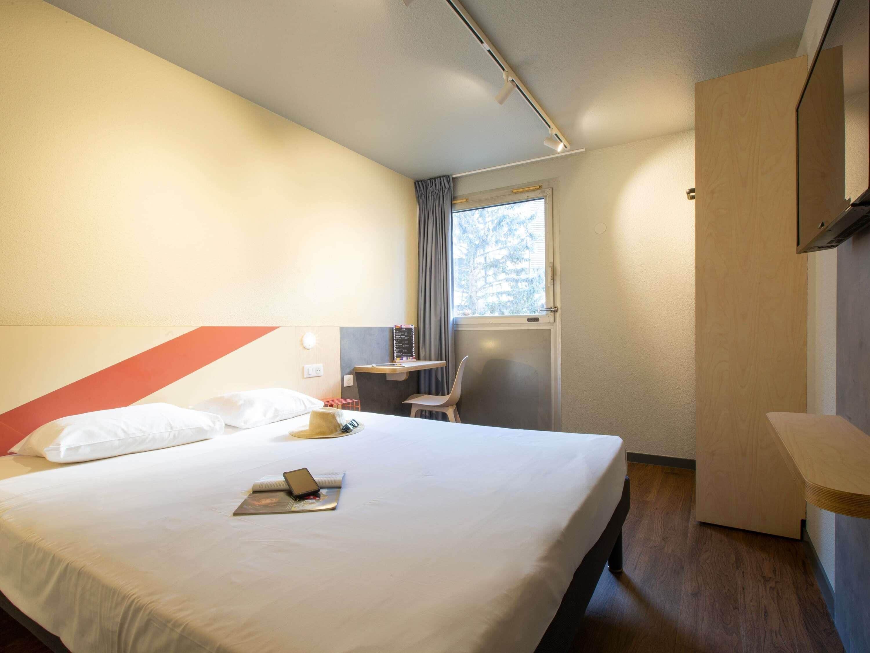 A hotel room with a bed, a desk, a chair, a window, and a mirror.