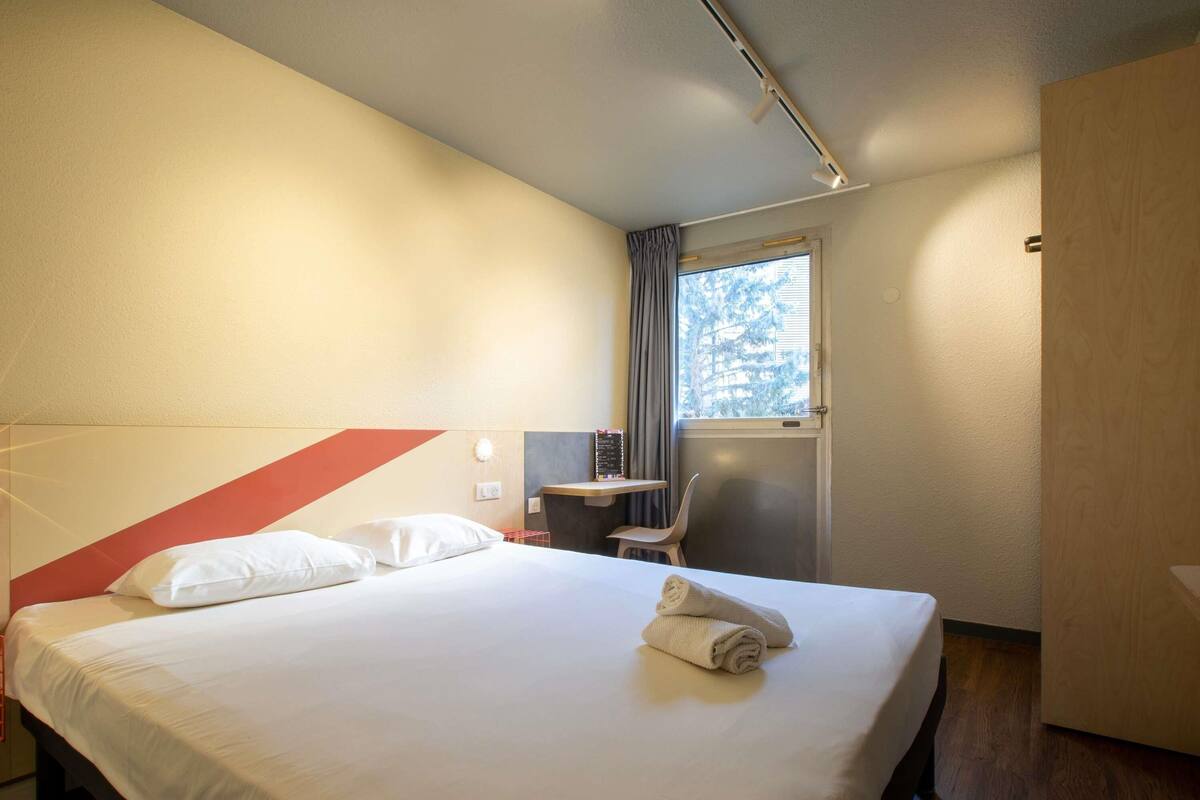 A hotel room with a bed, a desk, a chair, a window with curtains, and a door.