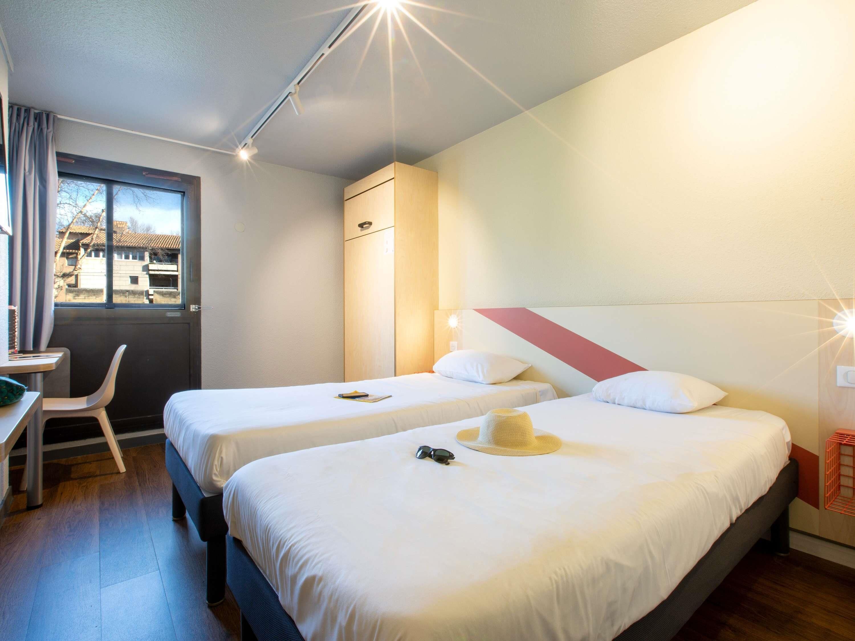 A hotel room with two beds, a desk, a chair, and a window with a view of buildings.