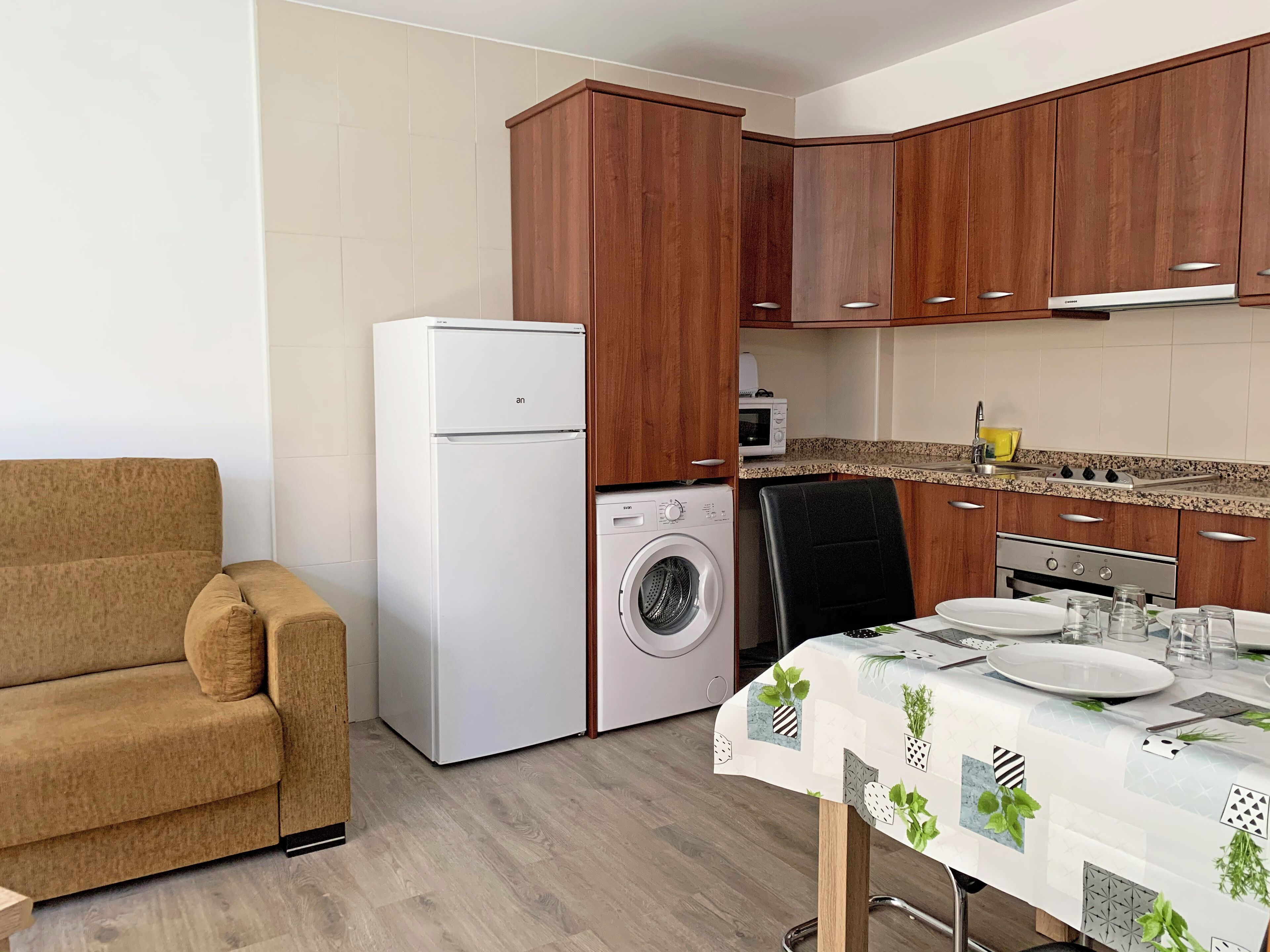 A compact kitchen with wooden cabinets, a white refrigerator, a washing machine, and a dining table set with plates and glasses.