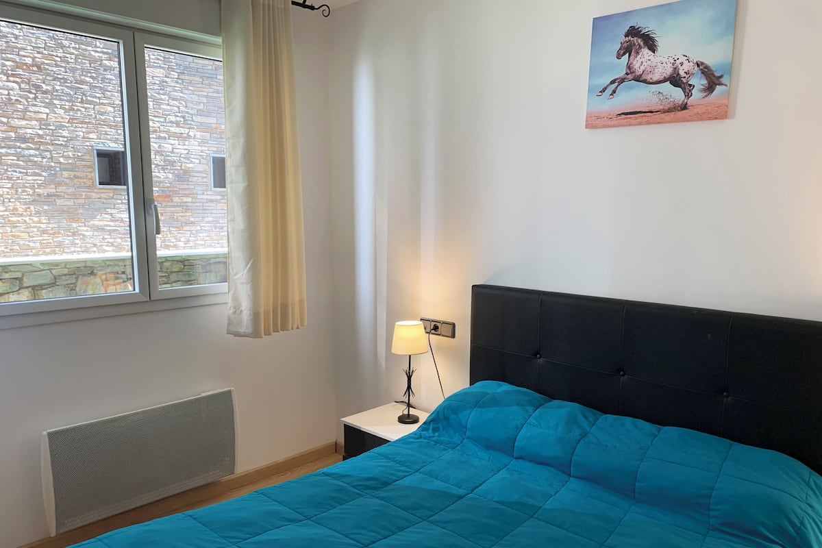 Quarto com cama grande, janela com cortinas, radiador, mesa de cabeceira com abajur e quadro de um cavalo na parede.