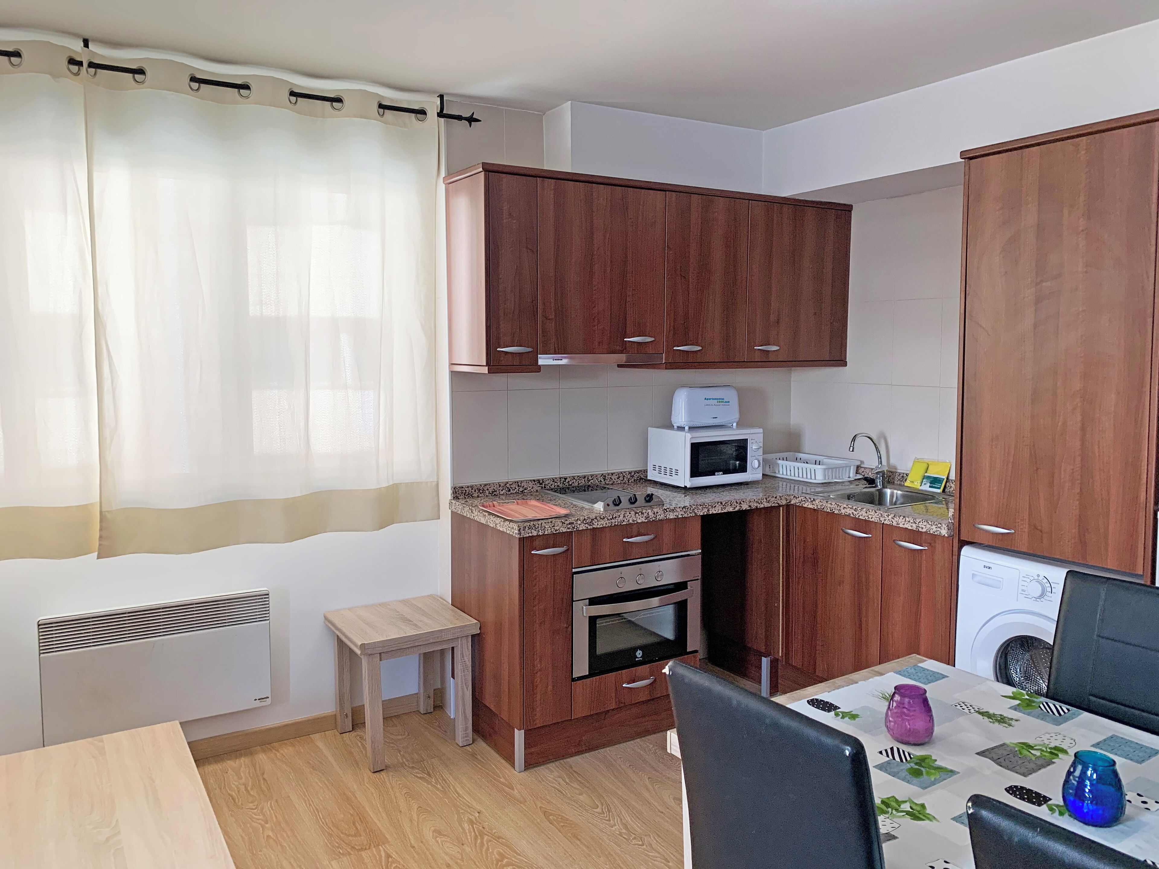 Una cocina compacta con armarios de madera, un microondas, un horno pequeño, un lavabo y una mesa con un mantel individual.