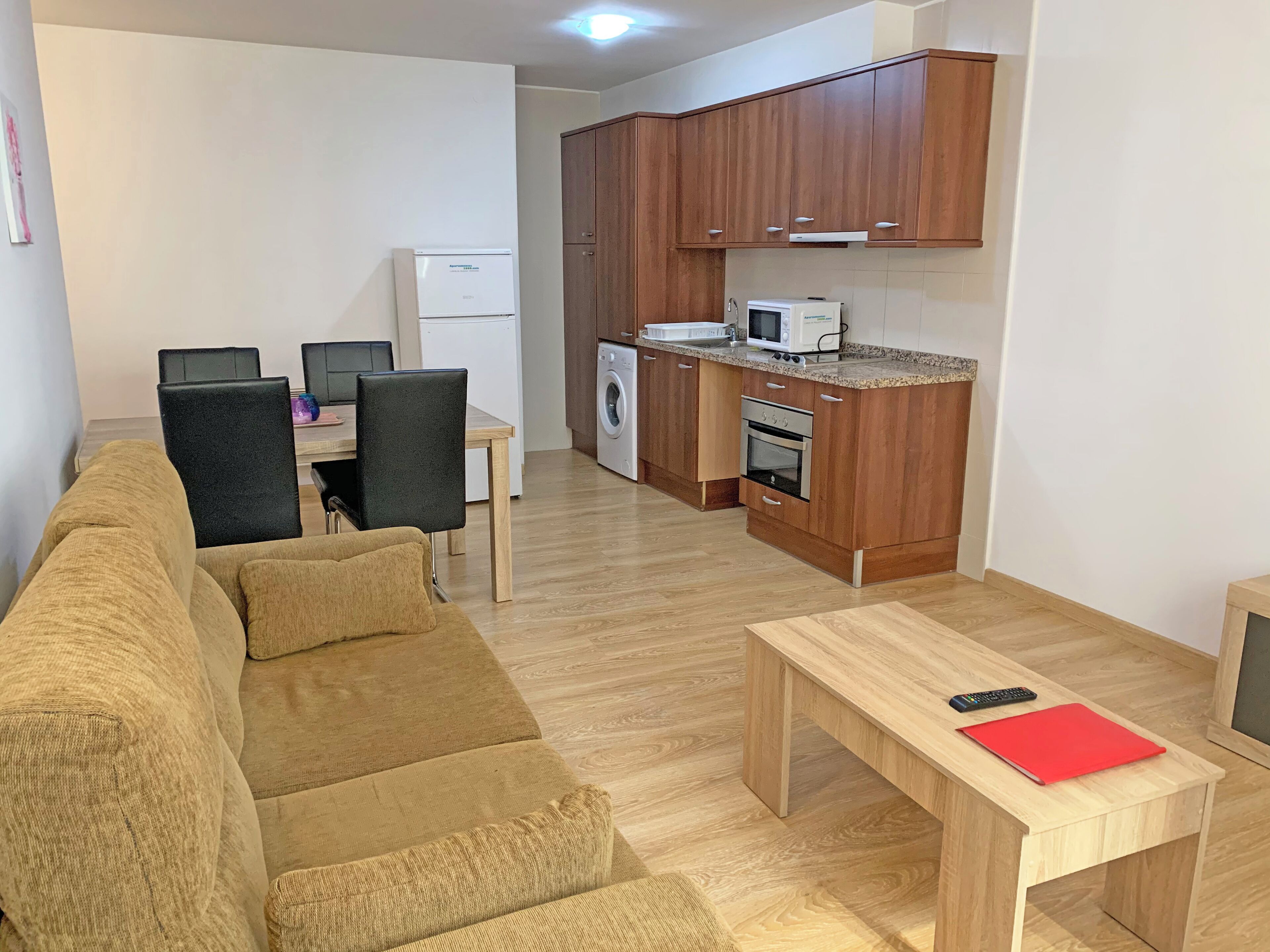 Una sala de estar con un sofá, una mesa de comedor con sillas, una cocina con armarios de madera, una lavadora y un horno.