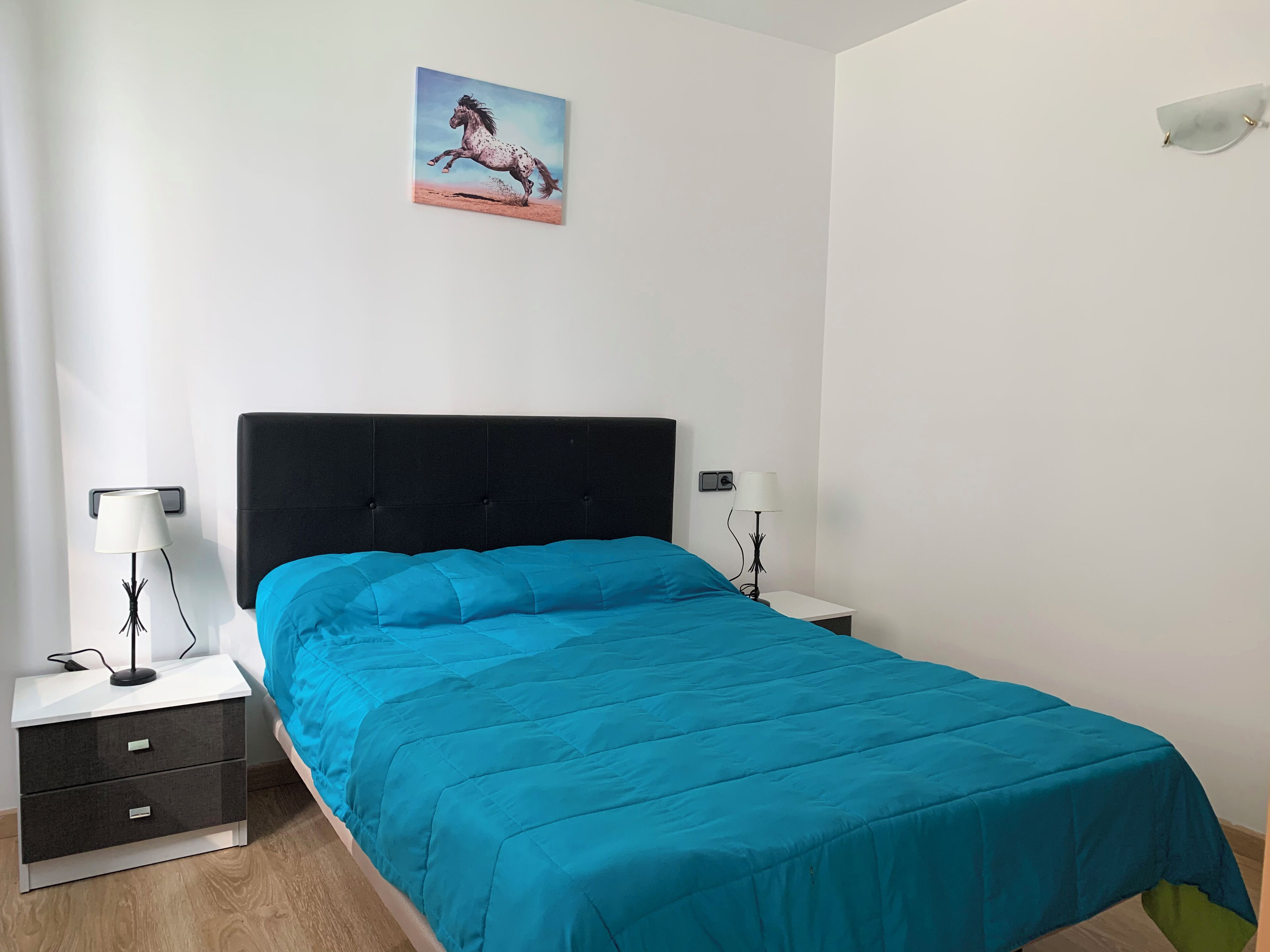 A bedroom with a black headboard, a blue bedspread, two bedside tables with lamps, and a wall-mounted painting.