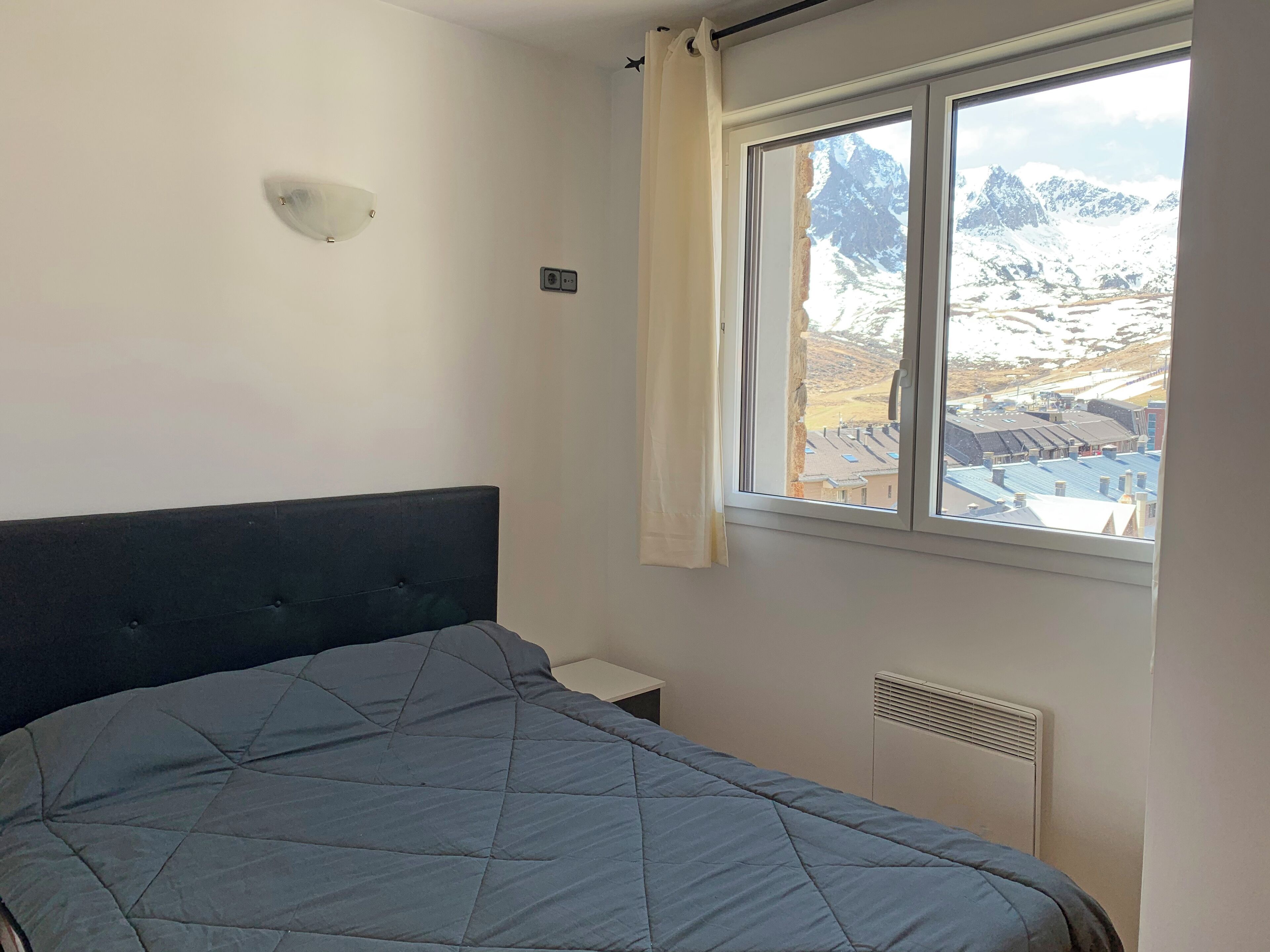Un dormitorio con cama, ventana con vistas a montañas nevadas, luz de pared y termostato.