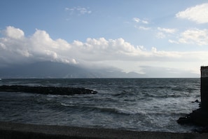 Paisaje costero con olas rompiendo contra rocas, cielo despejado y montañas a lo lejos.
