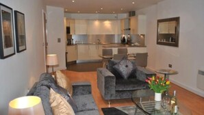A modern living room with a grey sofa, a glass coffee table, and a small dining area. - NG Apartments London (London)