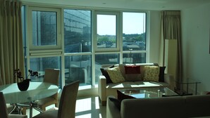 A modern living room with a glass coffee table, a sofa with patterned cushions, and a dining table with chairs. Large windows provide a view of the cityscape. - NG Apartments London (London)