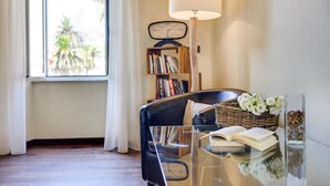 Een kamer met een lederen fauteuil, een glazen tafel, een lamp, een boekenkast en een raam met gordijnen.