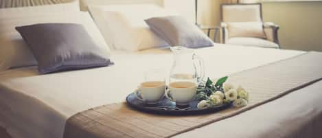 A neatly made bed with pillows, a tray with two cups of tea, a pitcher, and a bouquet of flowers.