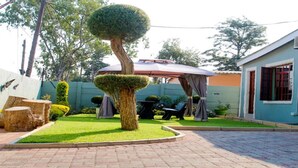 A courtyard with a neatly trimmed tree, a covered seating area, and a building with a blue facade. - Berry Bliss Guest House (Gaborone)