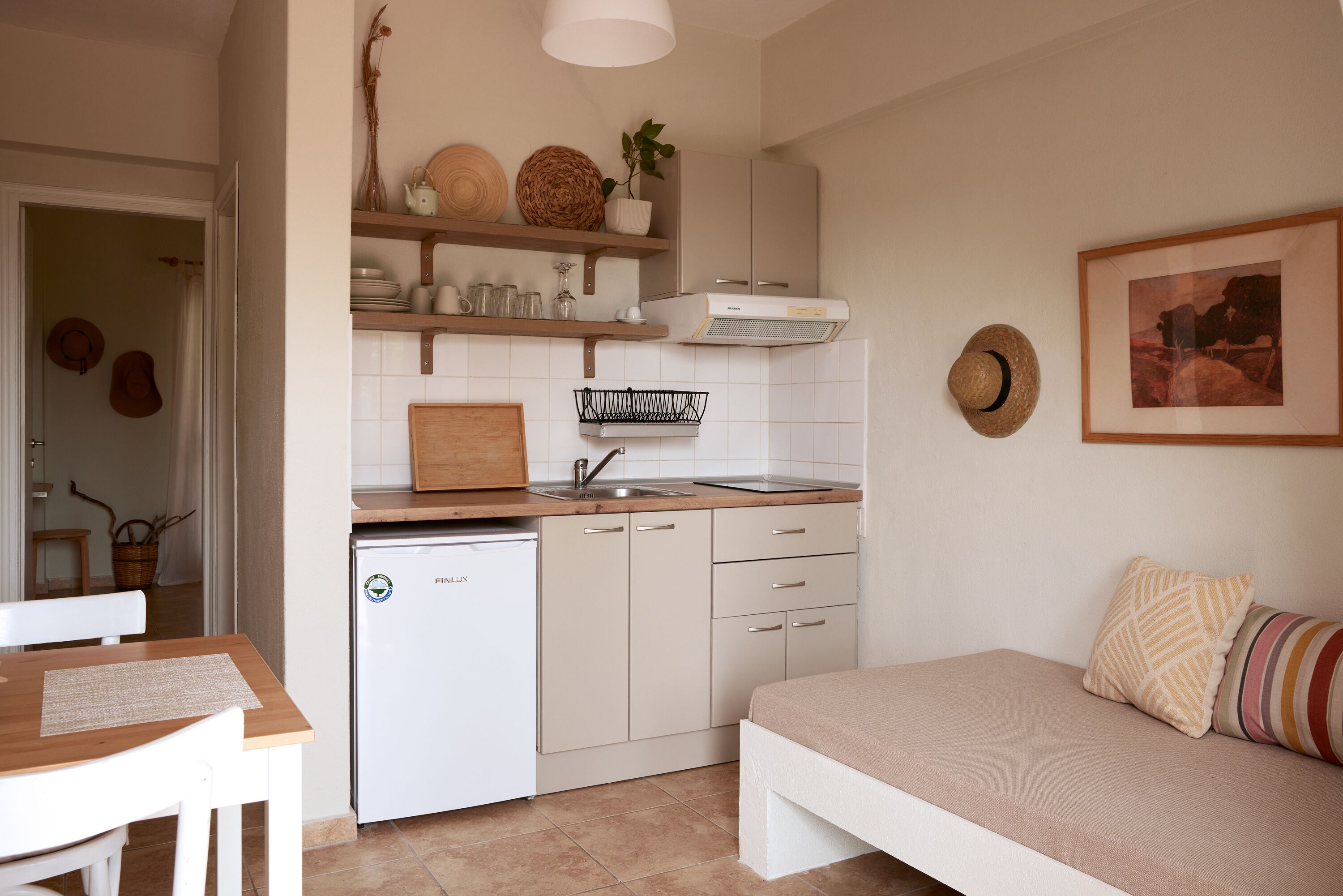 A compact kitchen with a sink, a small refrigerator, and a wooden countertop. There is a dining table with chairs and a bed with pillows.