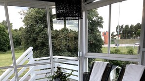A covered outdoor patio with a wooden table and chairs, a potted plant, and a view of the surrounding greenery and buildings. - Gävle Pendlarlägenhet (Gävle)