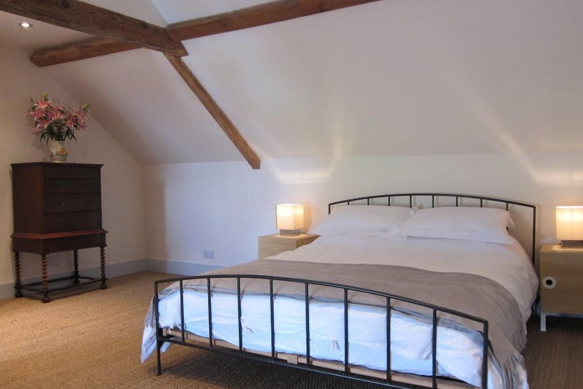A bedroom with a sloped ceiling, a metal bed frame, a wooden nightstand, a bedside table with a lamp, and a dresser with a vase of flowers.