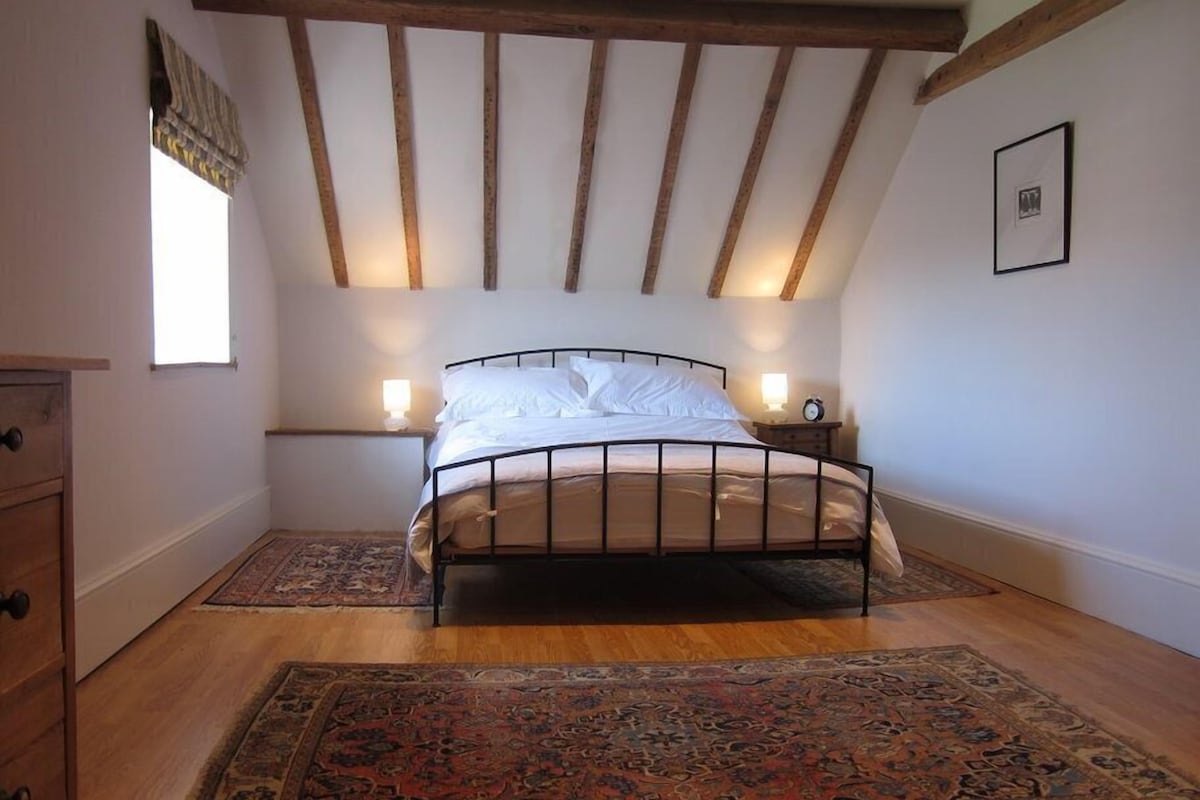A bedroom with a bed, wooden floor, a rug, a nightstand, and a window.