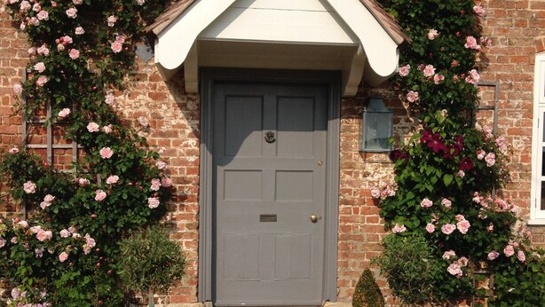 A brick building with a grey door, white trim, and a small window above the door. There are potted plants and a gravel pathway leading to the door. - Saul Farm (Gloucester)
