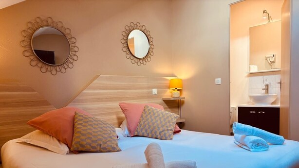 Ein Hotelzimmer mit einem Bett, Kissen, einem Nachttisch mit Lampe, einem Spiegel und einem durch eine offene Tür einsehbaren Badezimmer.