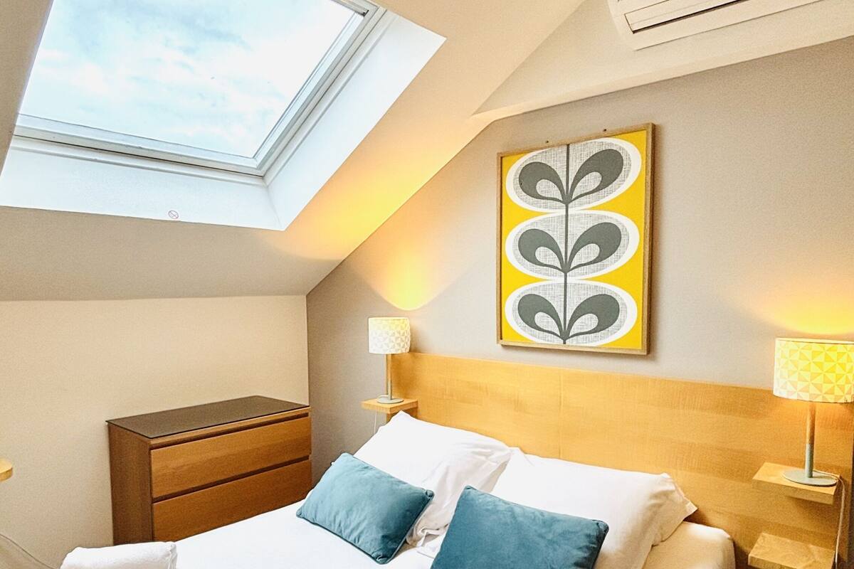 Une chambre d’hôtel comprenant un lit, une table de chevet, une lampe, une œuvre d’art murale et une lucarne.
