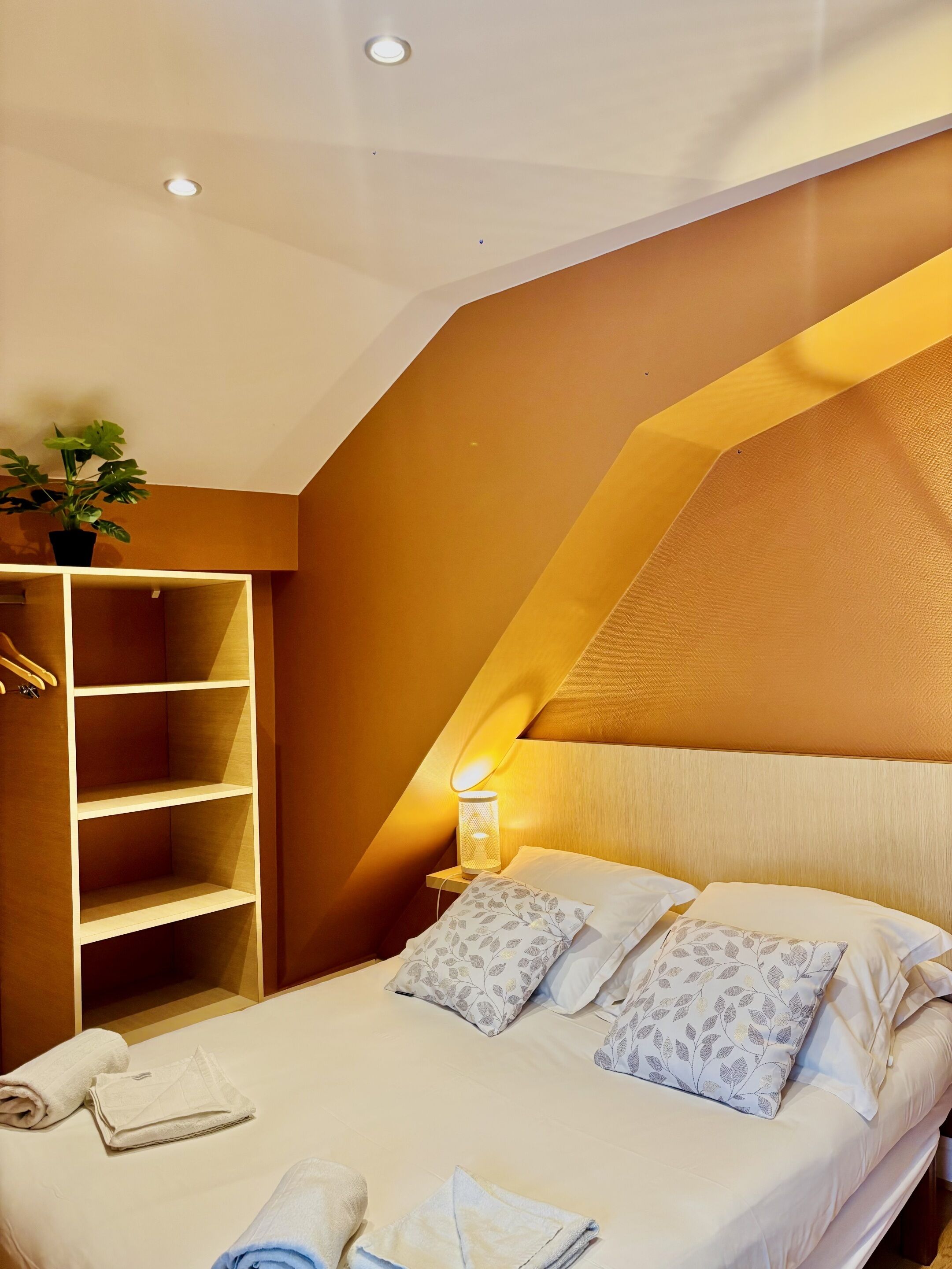 Un dormitorio con cama, almohadas, una mesita de noche con lámpara, una planta colgante y un armario empotrado.