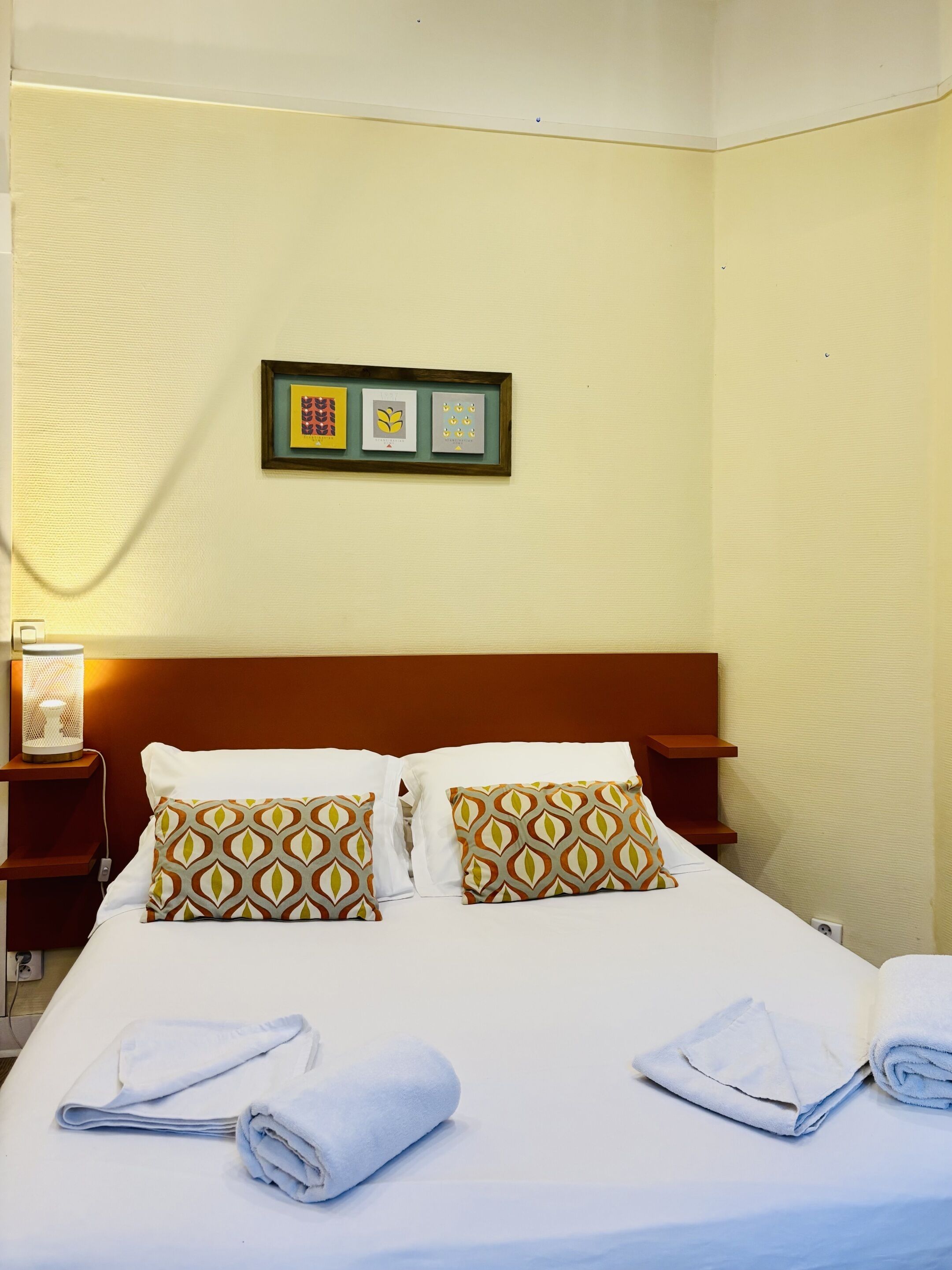 A hotel room with a bed, two pillows, a nightstand with a lamp, and a framed picture on the wall.