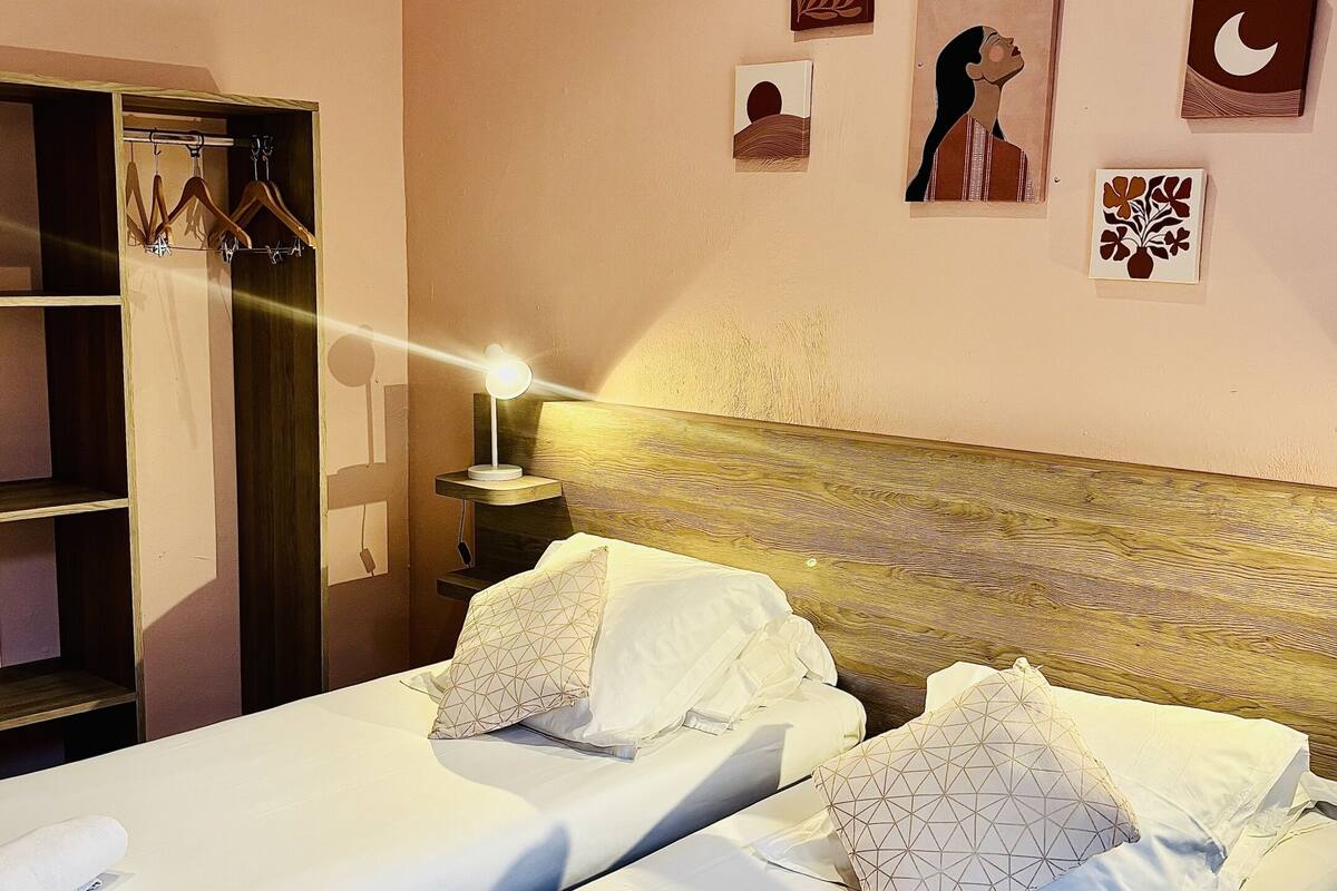 Une chambre d’hôtel avec deux lits, une armoire, une table de chevet avec une lampe et des œuvres d’art accrochées aux murs.