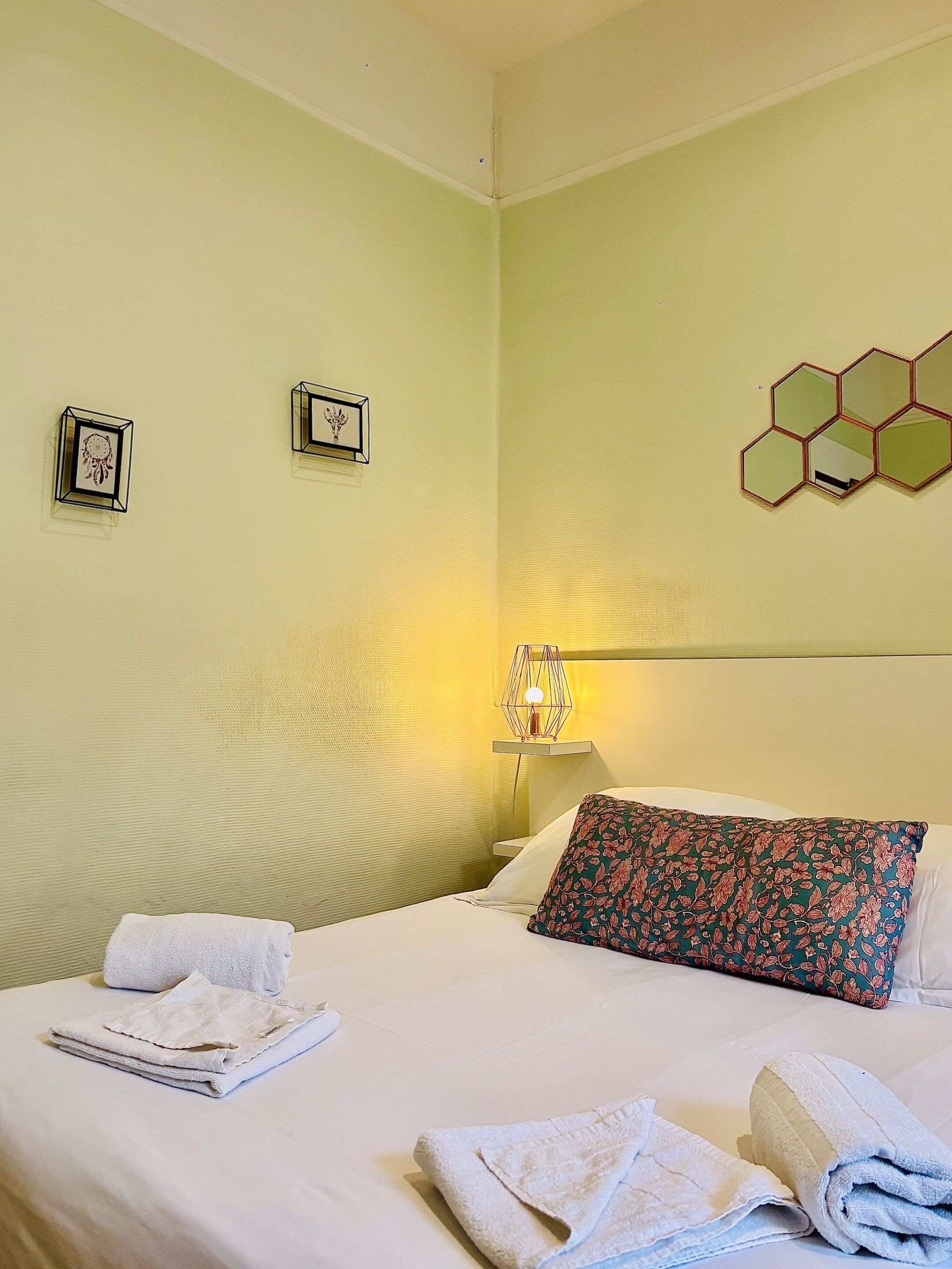 Quarto de hotel com uma cama, dois quadros pendurados na parede, um espelho hexagonal e uma luminária de cabeceira.