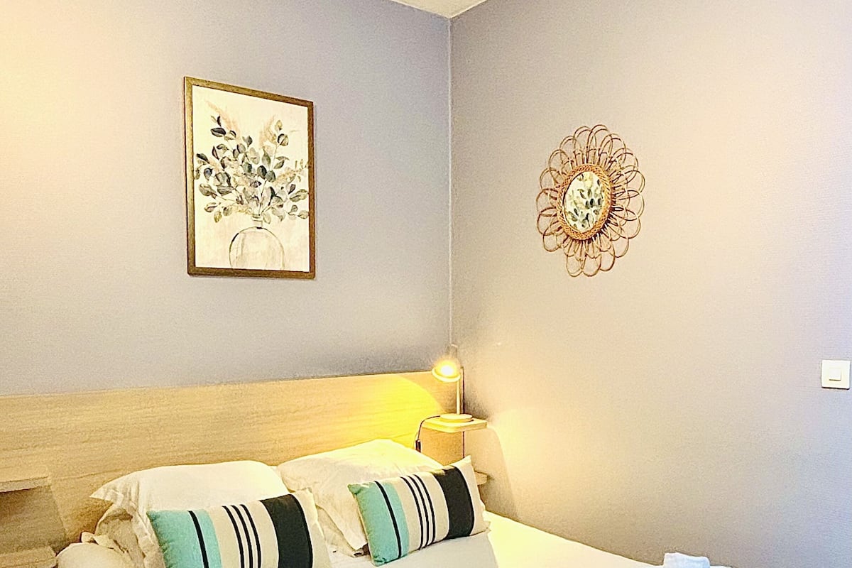 Un lit bien fait, avec des oreillers et des serviettes, une lampe de chevet et une estampe botanique encadrée au mur.