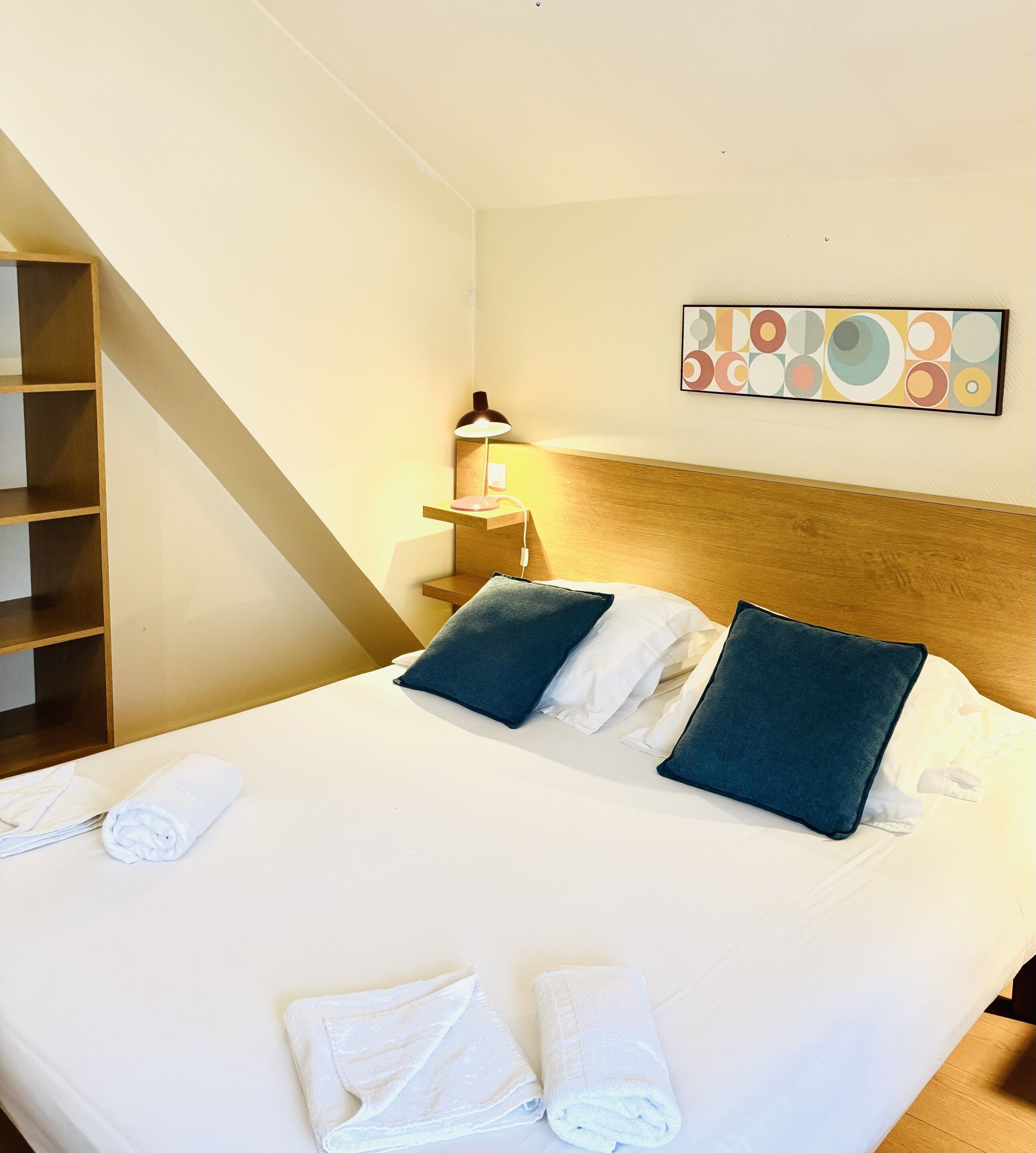 Un lit bien fait, avec des oreillers et des serviettes enroulées, une lampe de chevet, une tête de lit en bois et une œuvre d’art accrochée au mur.