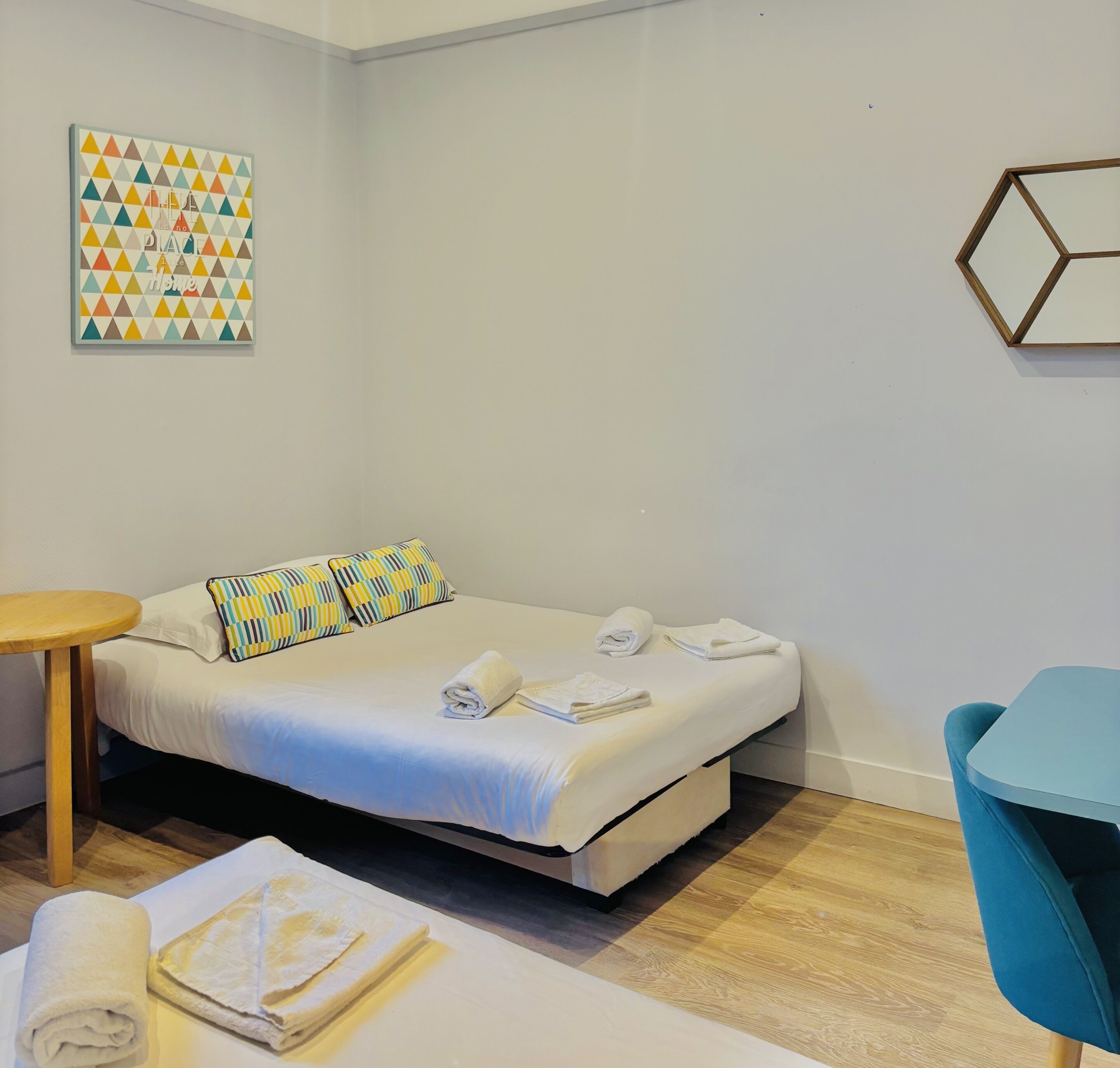 Un lit bien fait, avec des oreillers, une petite table ronde, une chaise bleue et un élément décoratif géométrique sur le mur.