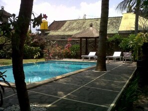 A swimming pool with a tiled deck, surrounded by palm trees and a thatched-roof structure. - Padang Lovina Sea Side Cottage (Buleleng)