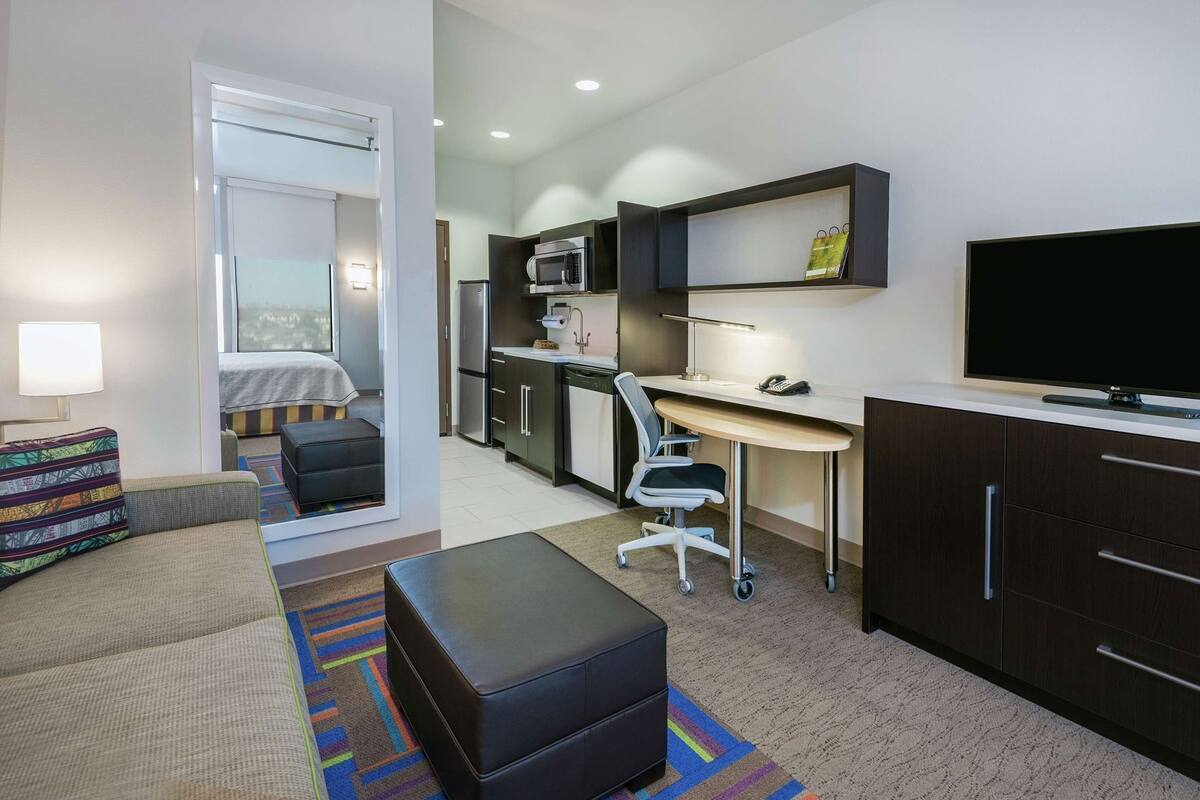 A compact hotel room with a kitchenette, a desk, a television, a sofa, and a small window.