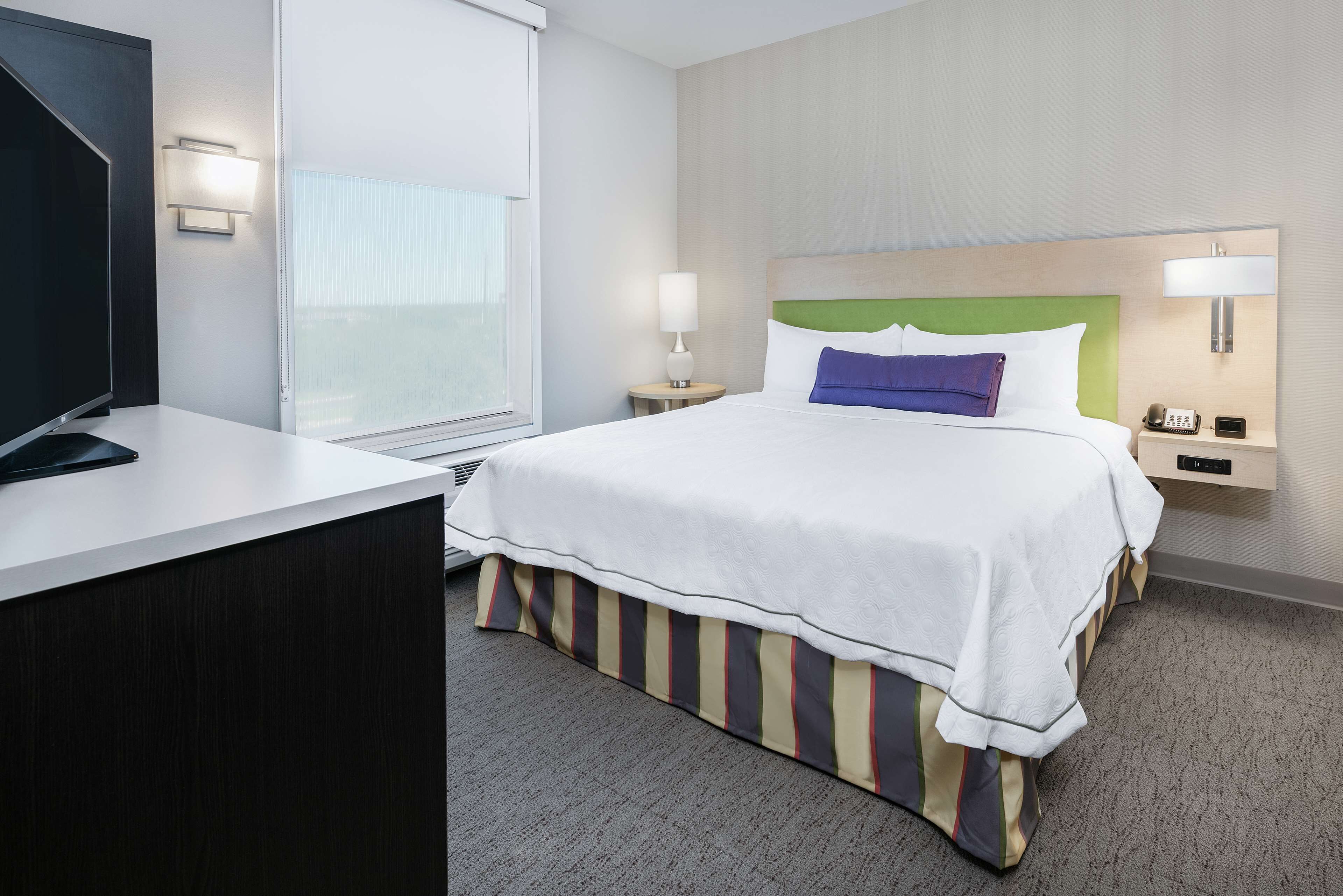 A hotel room with a bed, bedside tables, a TV, and a window.